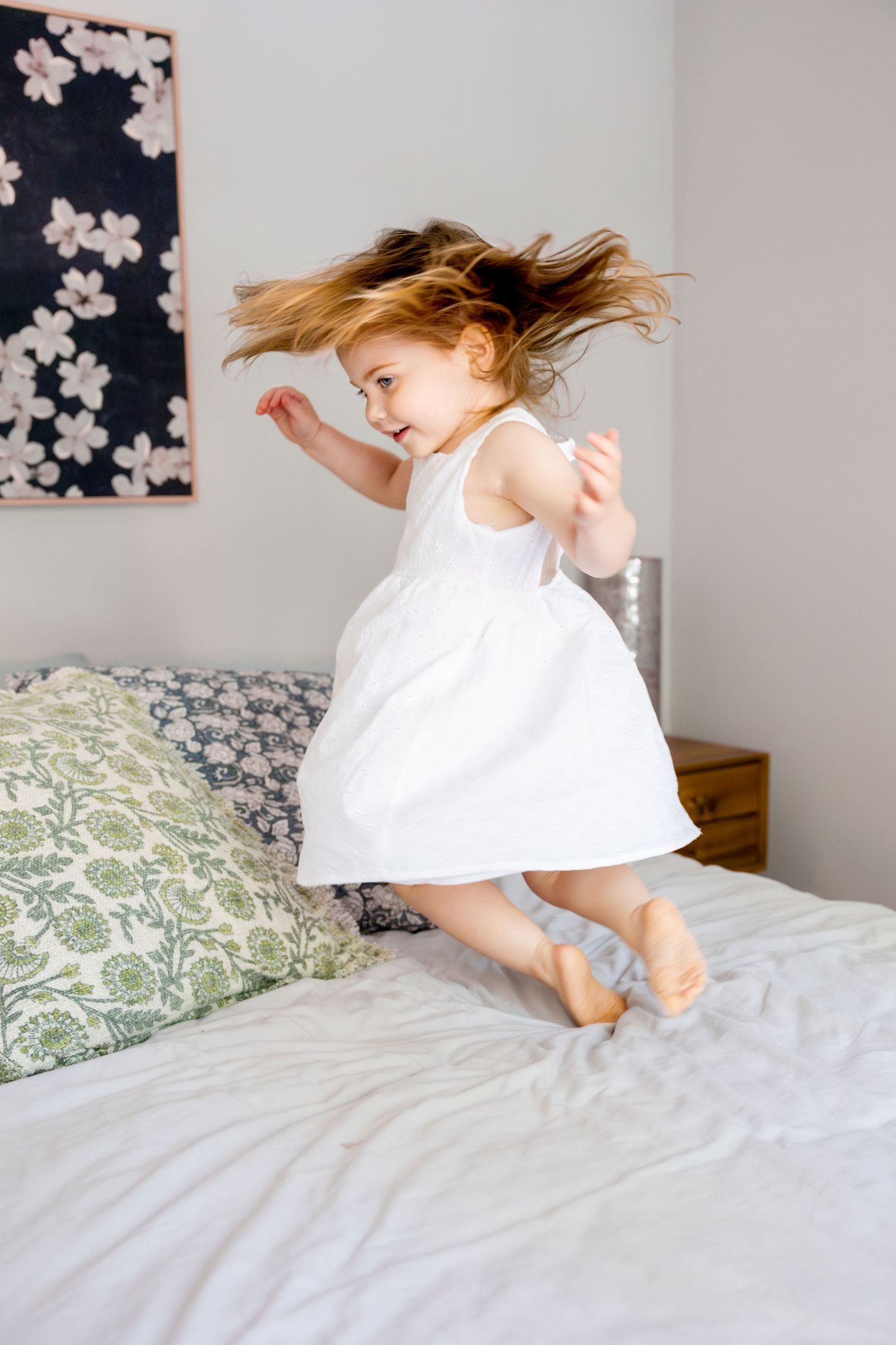 A child in a white dress joyfully jumping on a bed with floral pillows and wall art in the background.