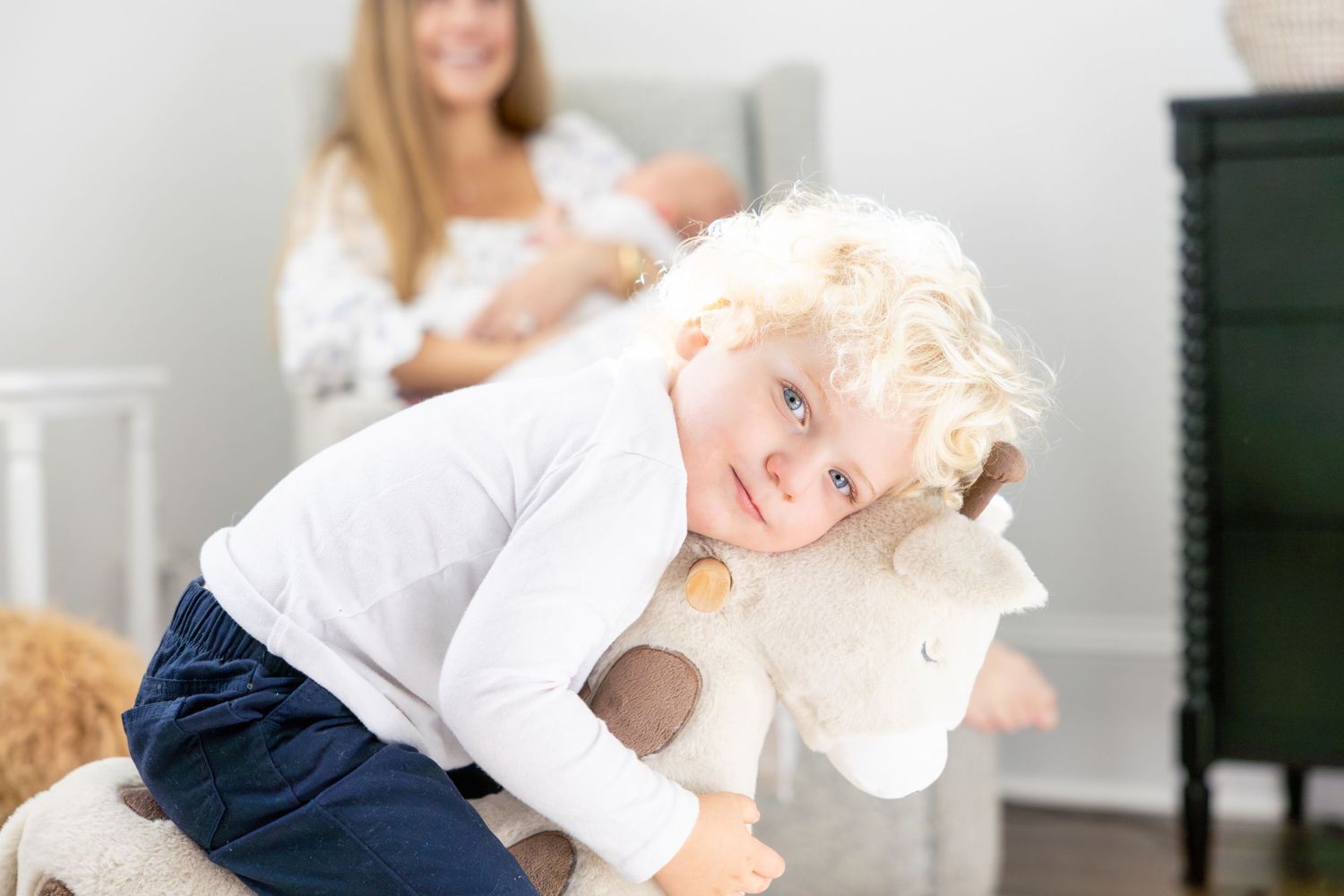 Toddler plays with stuffed animal while mother holds baby in background.