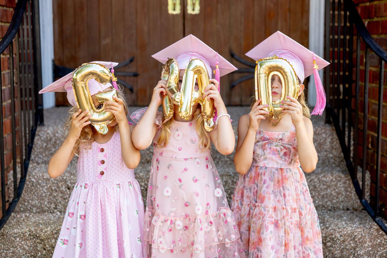 Three preschool graduates in pink dresses and graduation caps hide behind gold letter balloons that spell out GRAD.