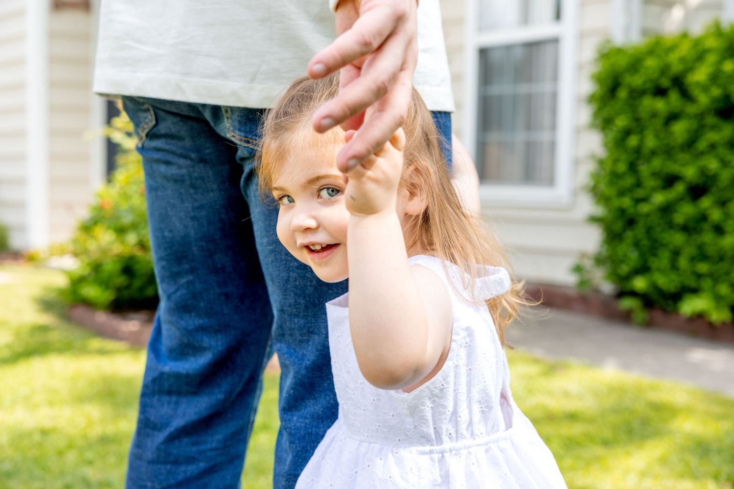 Child in white dress reaching up while playing outdoors near a house.
