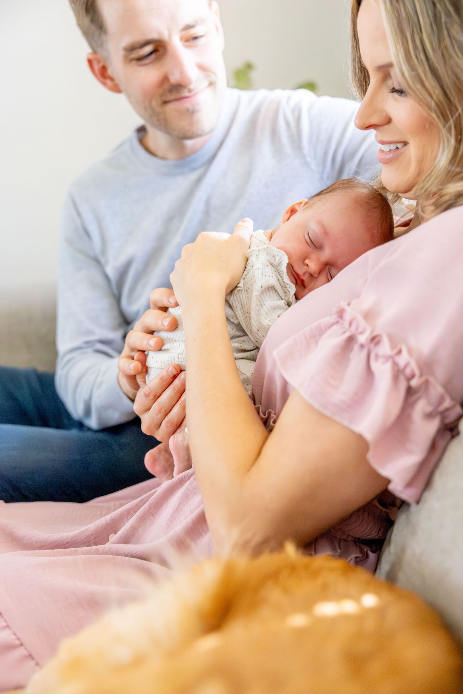 Couple on couch sharing tender moment while holding their sleeping newborn.