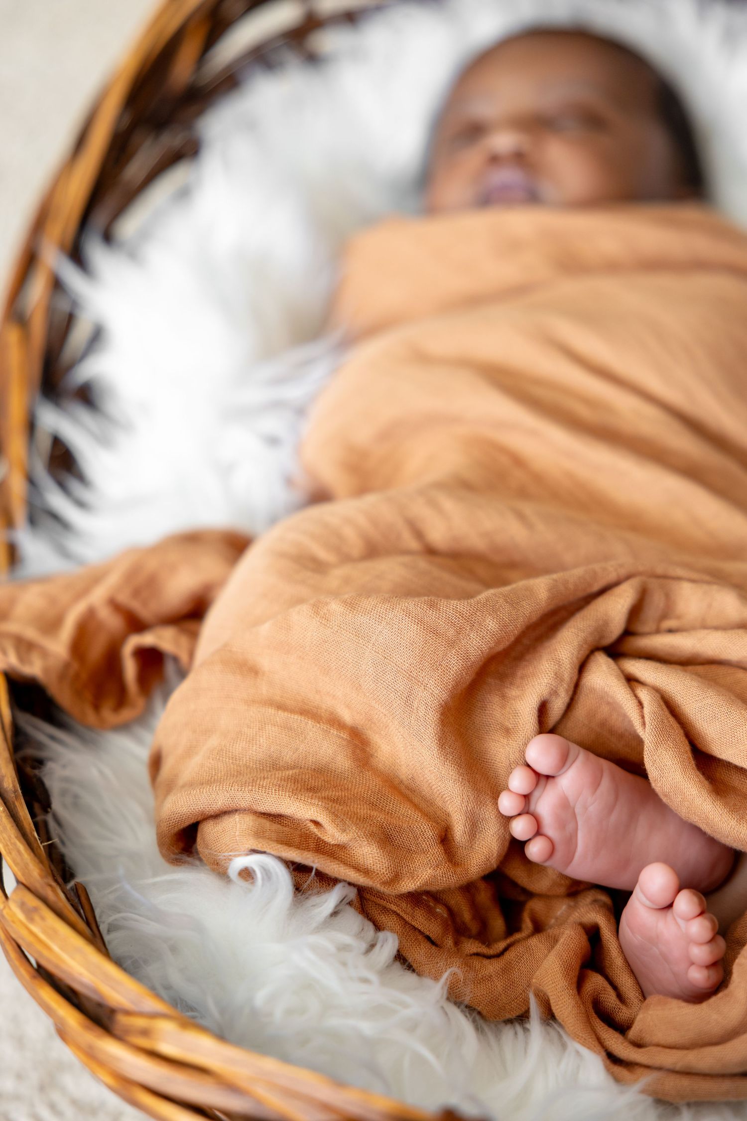 Tiny feet peek out from orange swaddle blanket in white fur-lined basket.