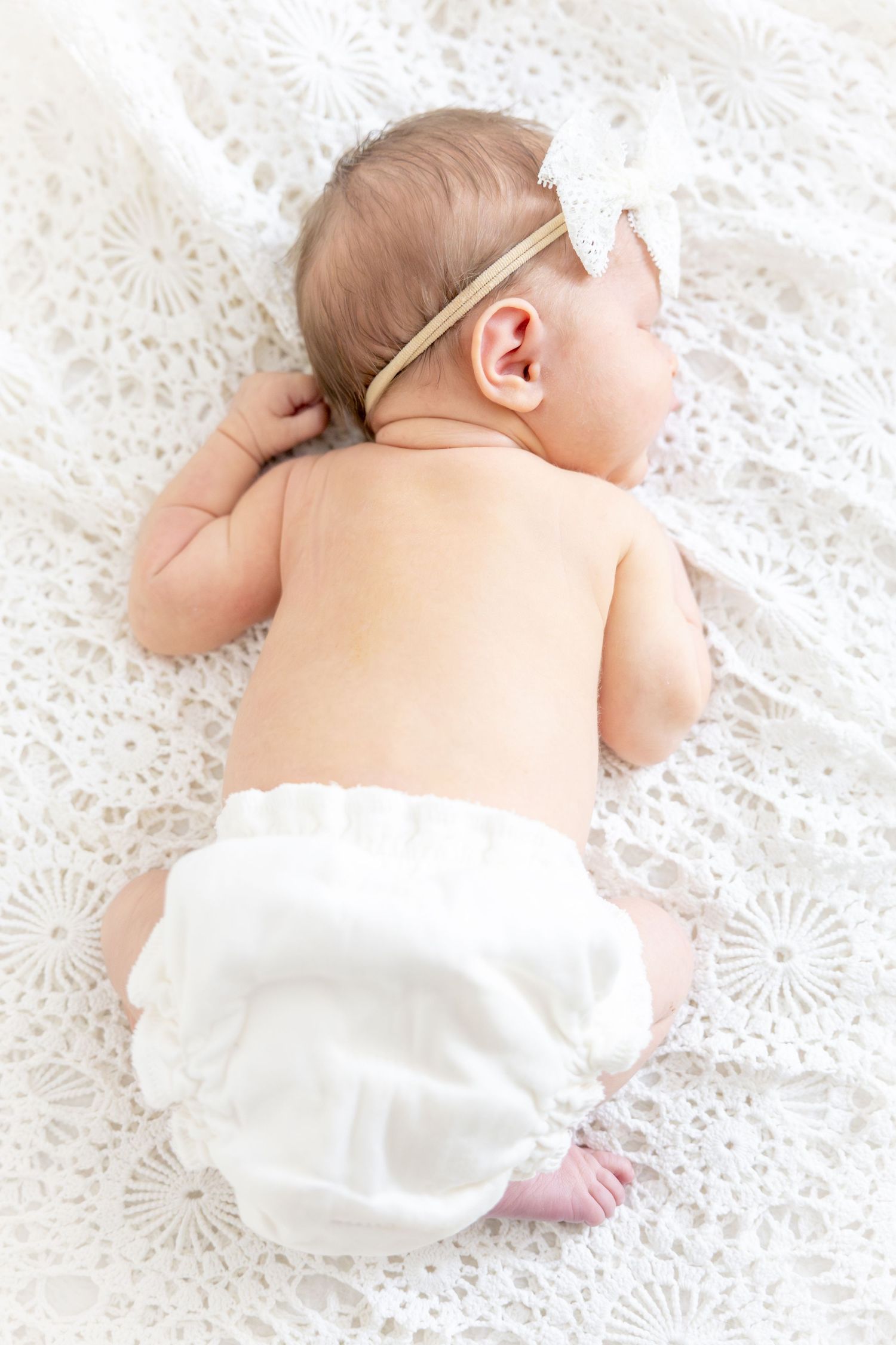 Baby laying on white lace blanket wearing diaper and delicate headband.