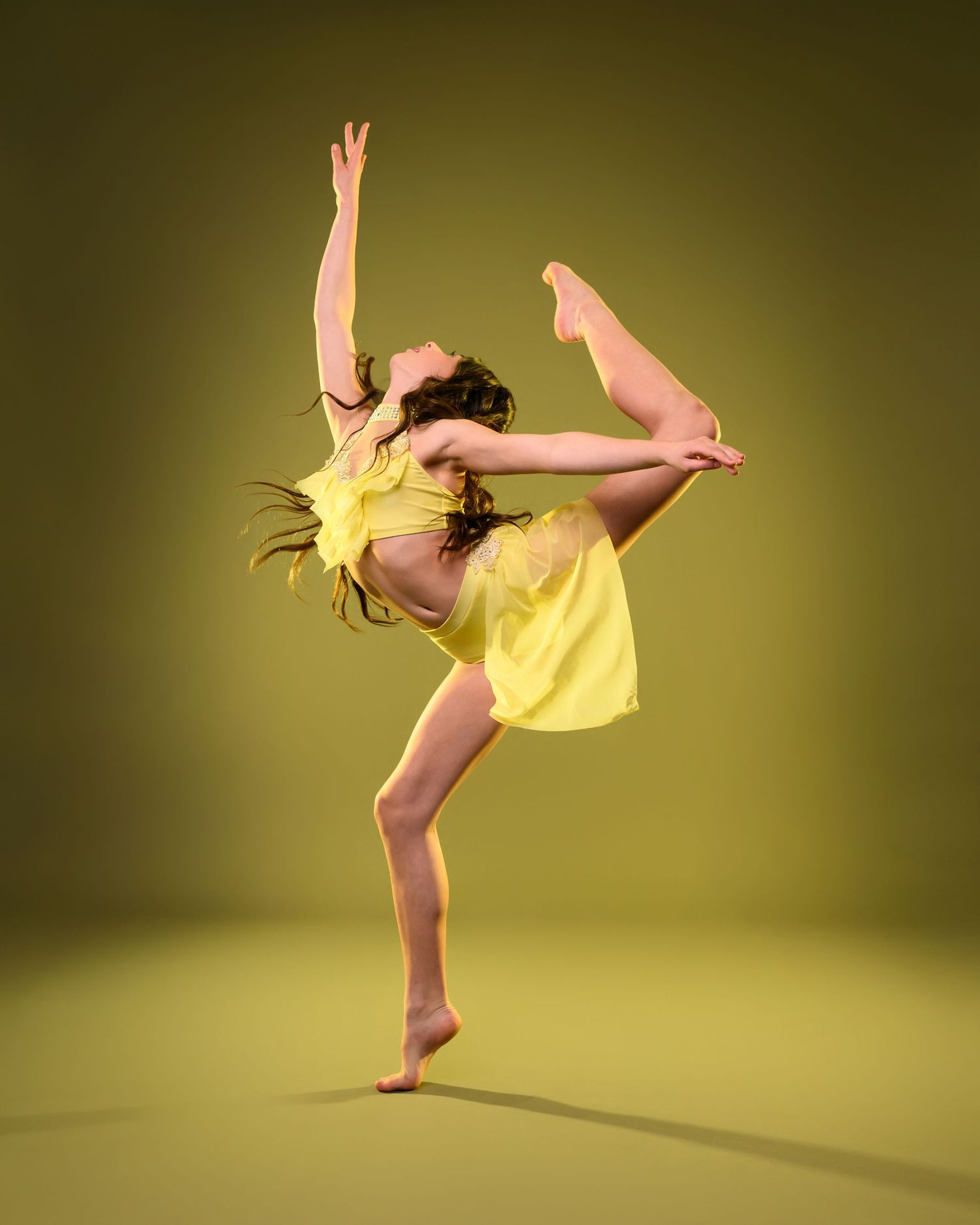 A dancer in a yellow dress leaps gracefully against a yellow backdrop, extending one leg high while reaching arms upward.