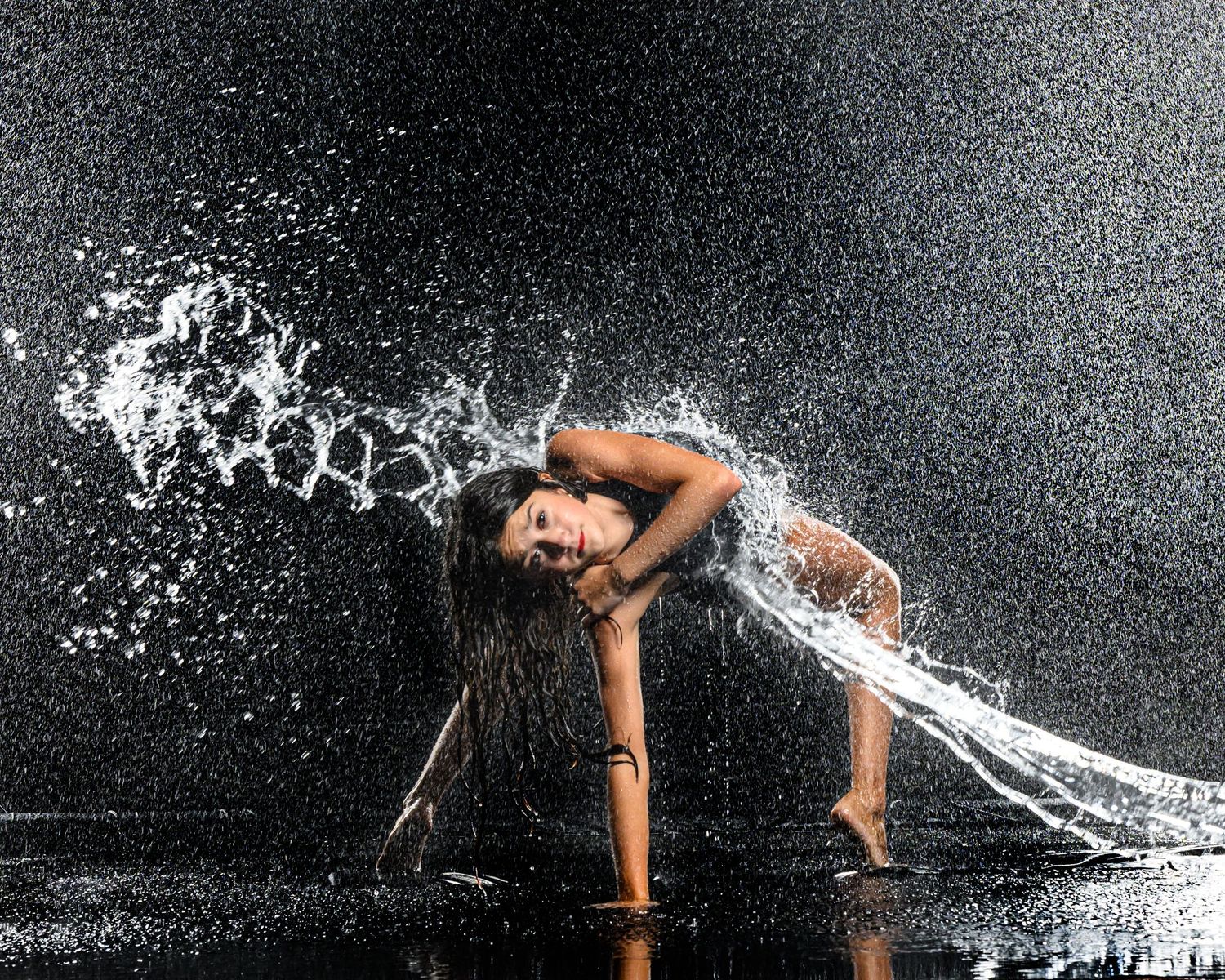 A woman dances dramatically in splashing water against a dark background, her wet hair creating an arc of droplets.