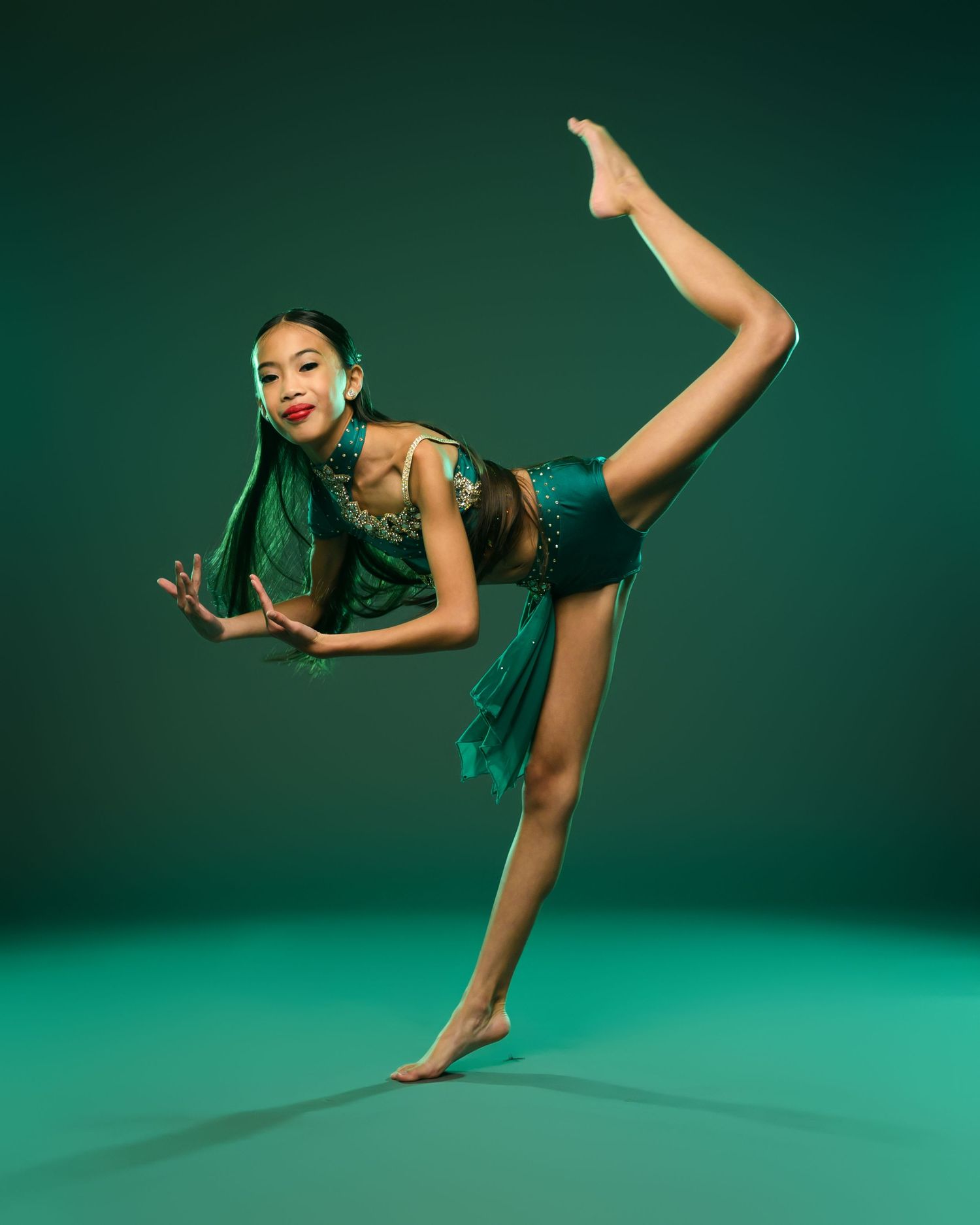 Dancer in emerald green costume performs arabesque pose against green studio background.