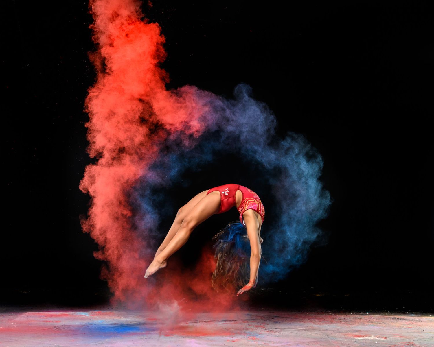 Gymnast performing a back handspring through colorful smoke effects against dark background.