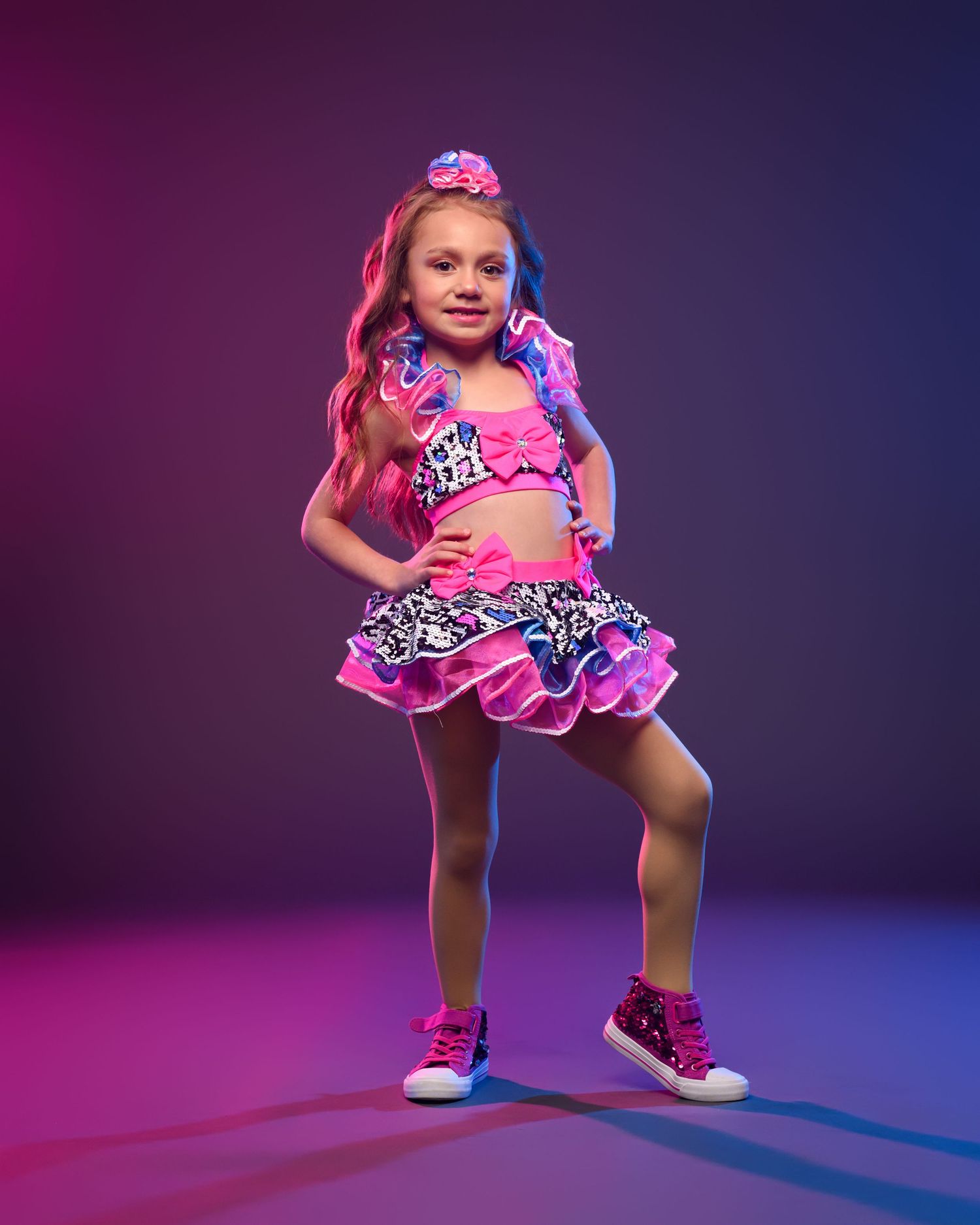 Young dancer strikes pose in pink ruffled dance costume with colorful lighting effects.