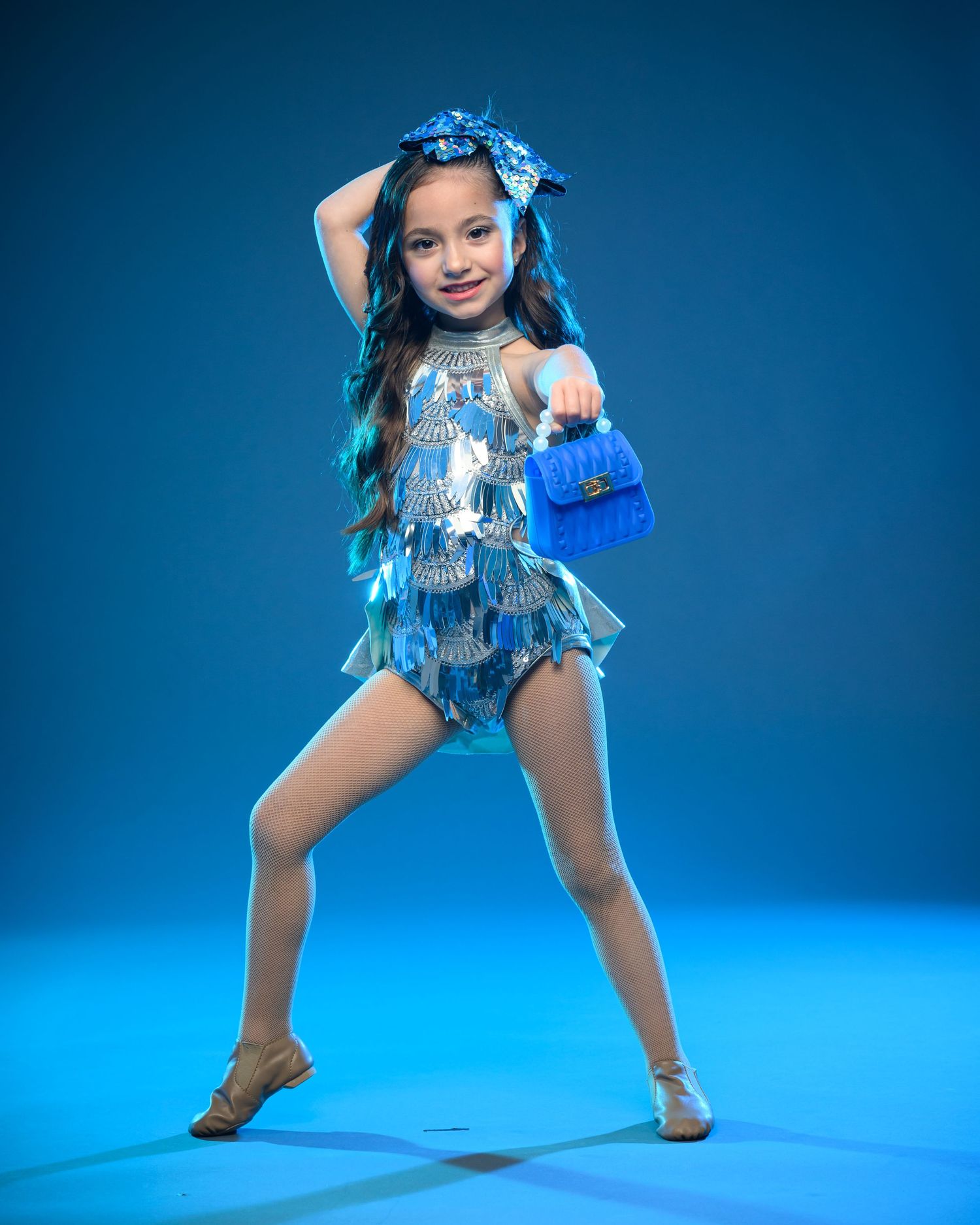 Young dancer in sparkly blue costume strikes confident pose against vibrant blue background.