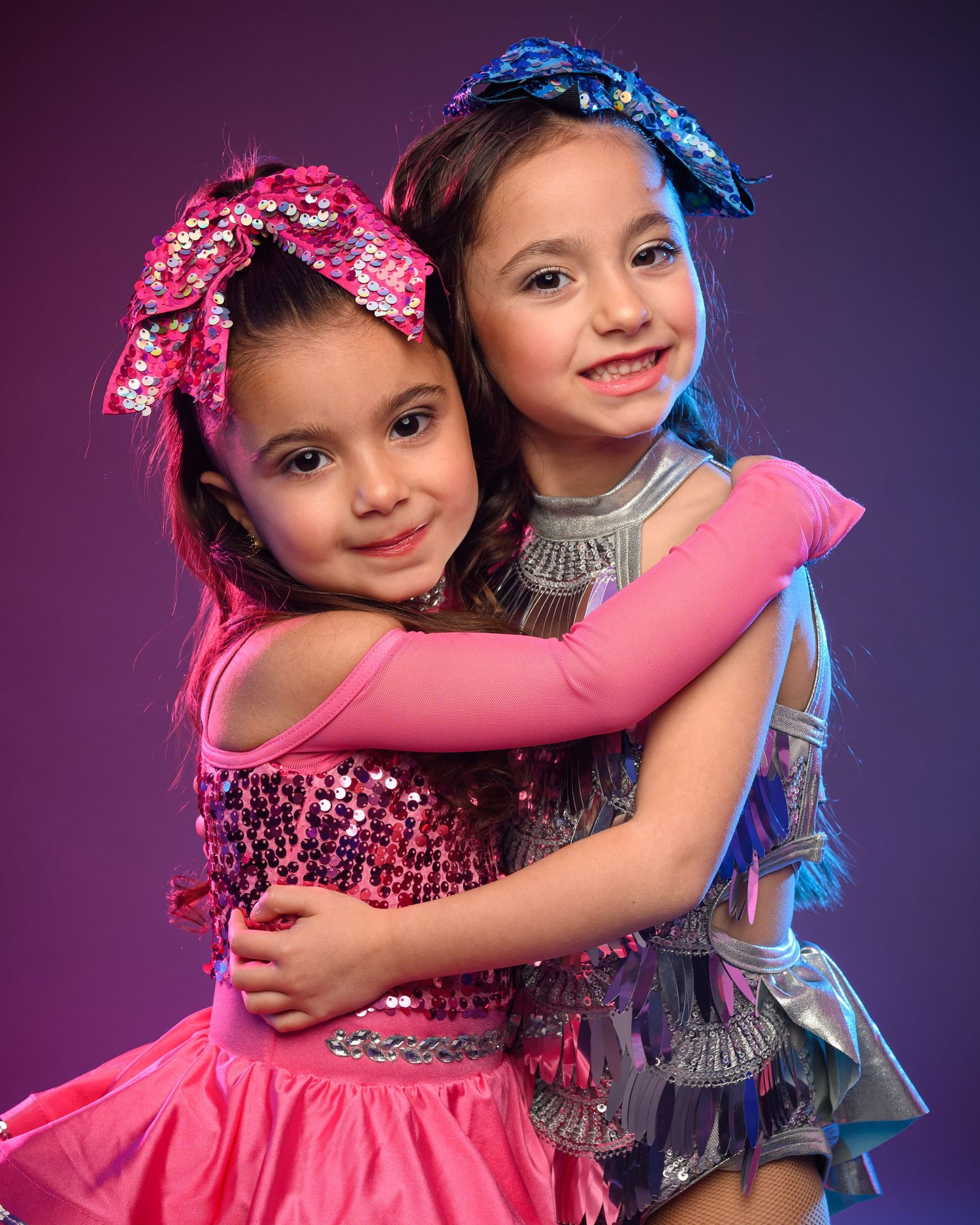 Two dancers in contrasting pink and blue costumes embrace in a friendly performance pose.