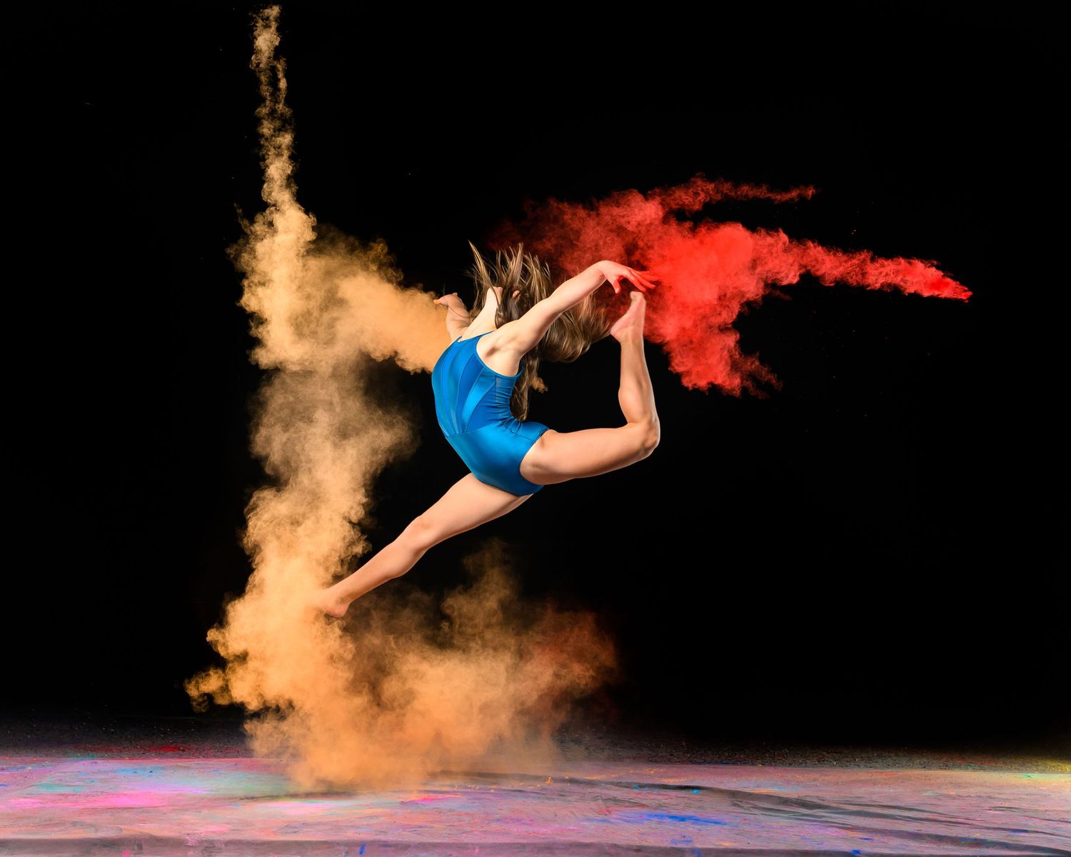 A silhouetted gymnast leaps gracefully through clouds of colorful powder against a dark background.