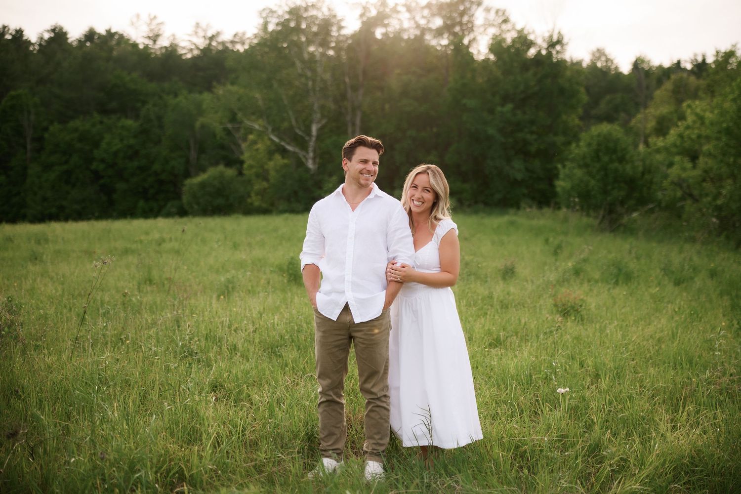 Couple in white outfits smiling together in a sunny green meadow with trees in the background.