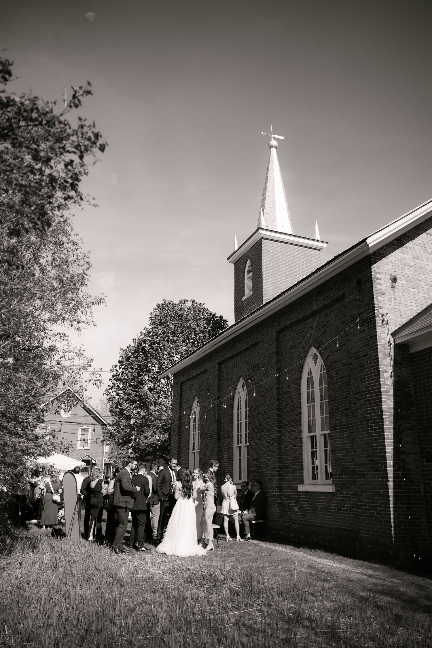 Black and white photo of a wedding party gathered outside a historic brick church with a steeple on a sunny day.