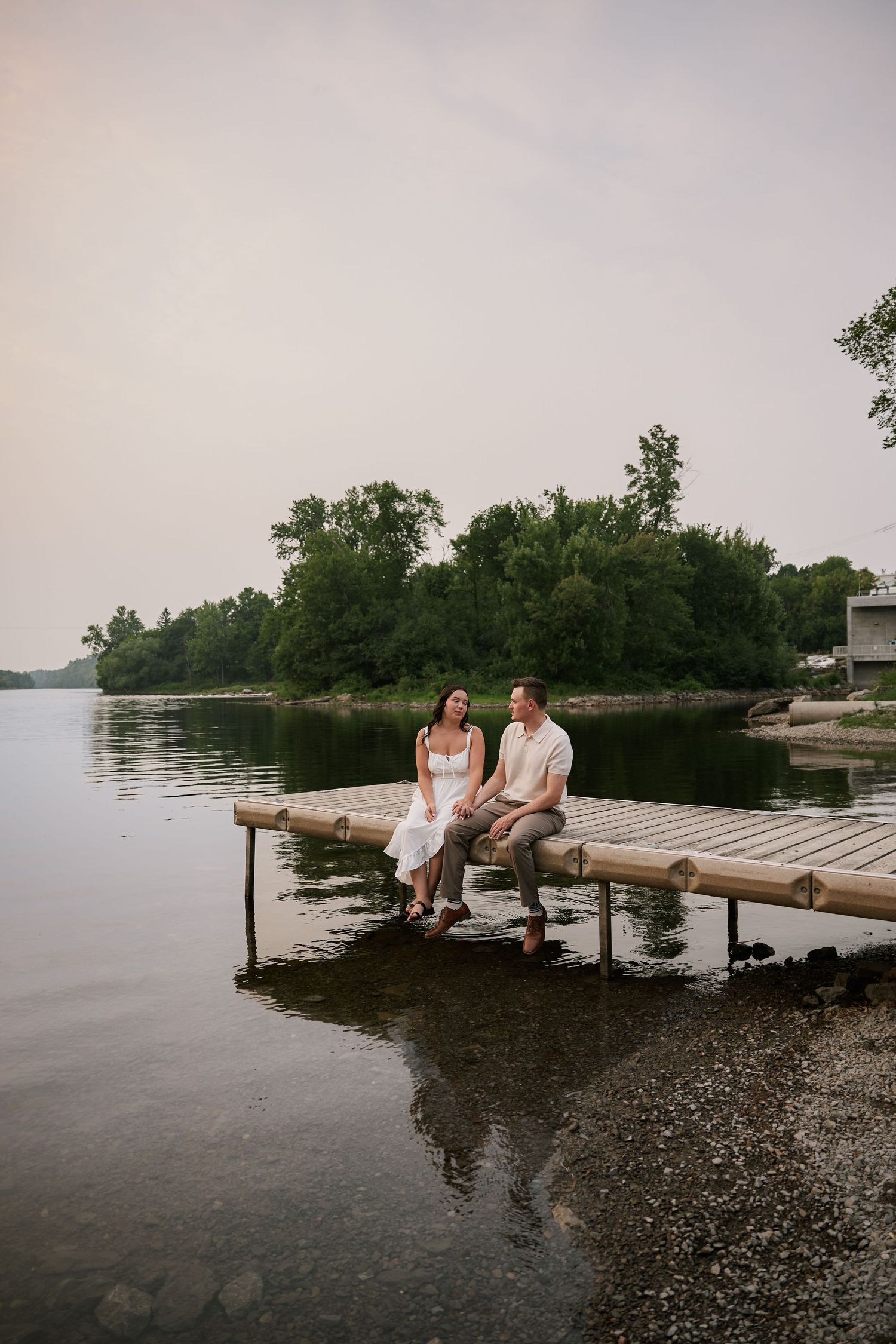Couple in wedding attire sitting together on a wooden dock by a calm lake surrounded by lush green trees.