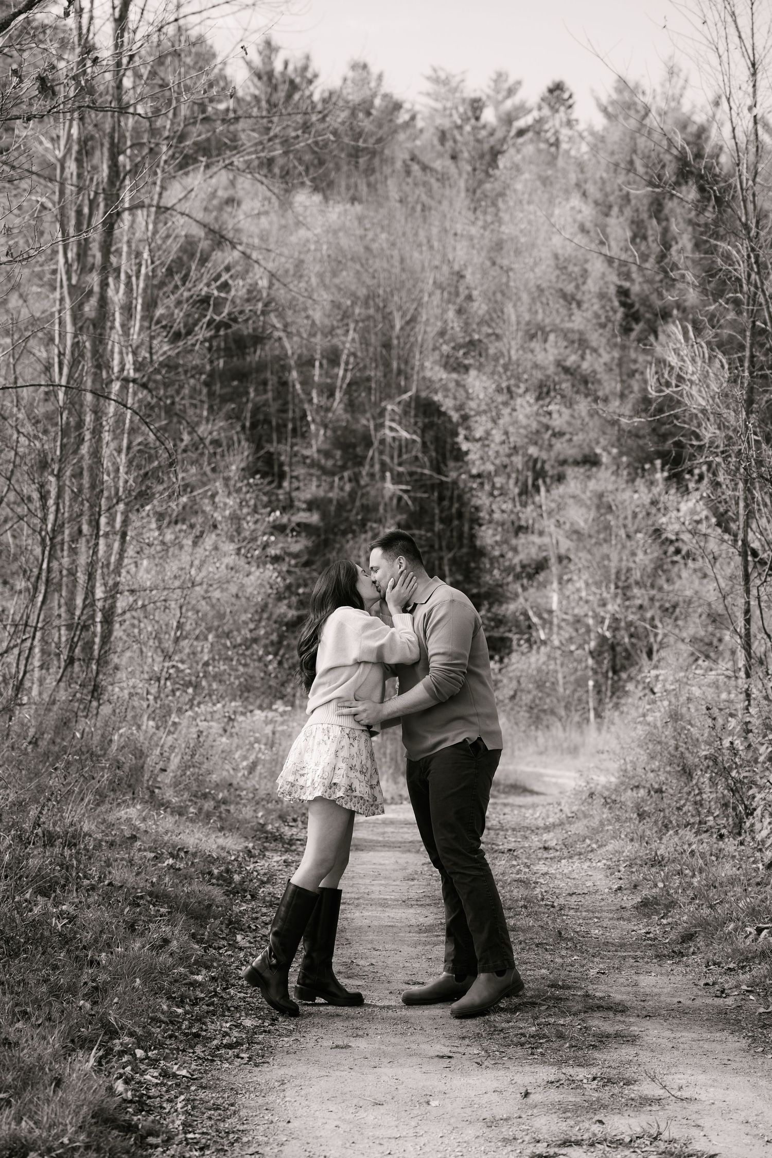 Black and white photo of a couple kissing on a forest path surrounded by tall trees.