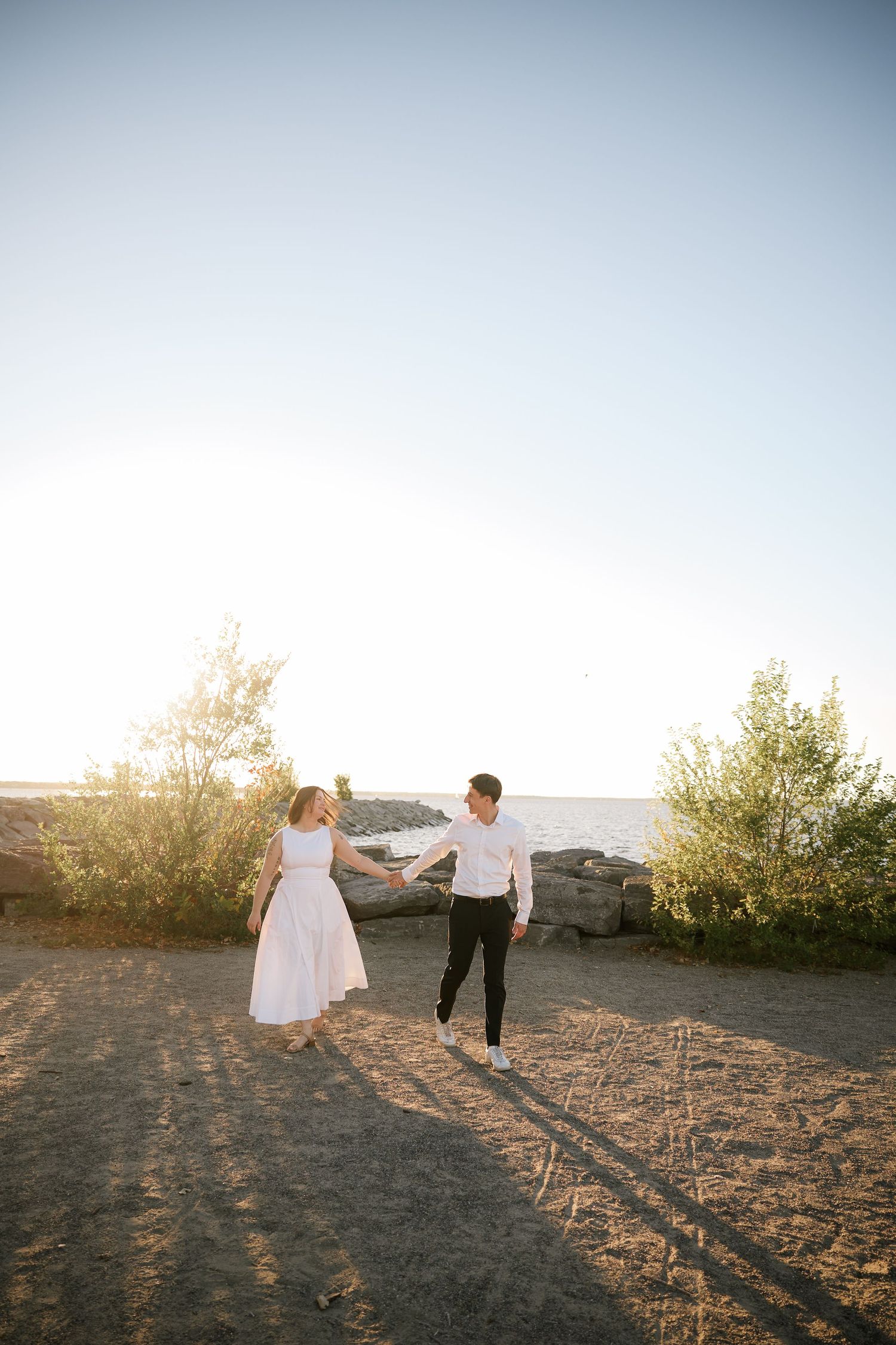 Couple holding hands walking near rocky shoreline at golden hour, woman in white dress, man in black pants and white shirt.