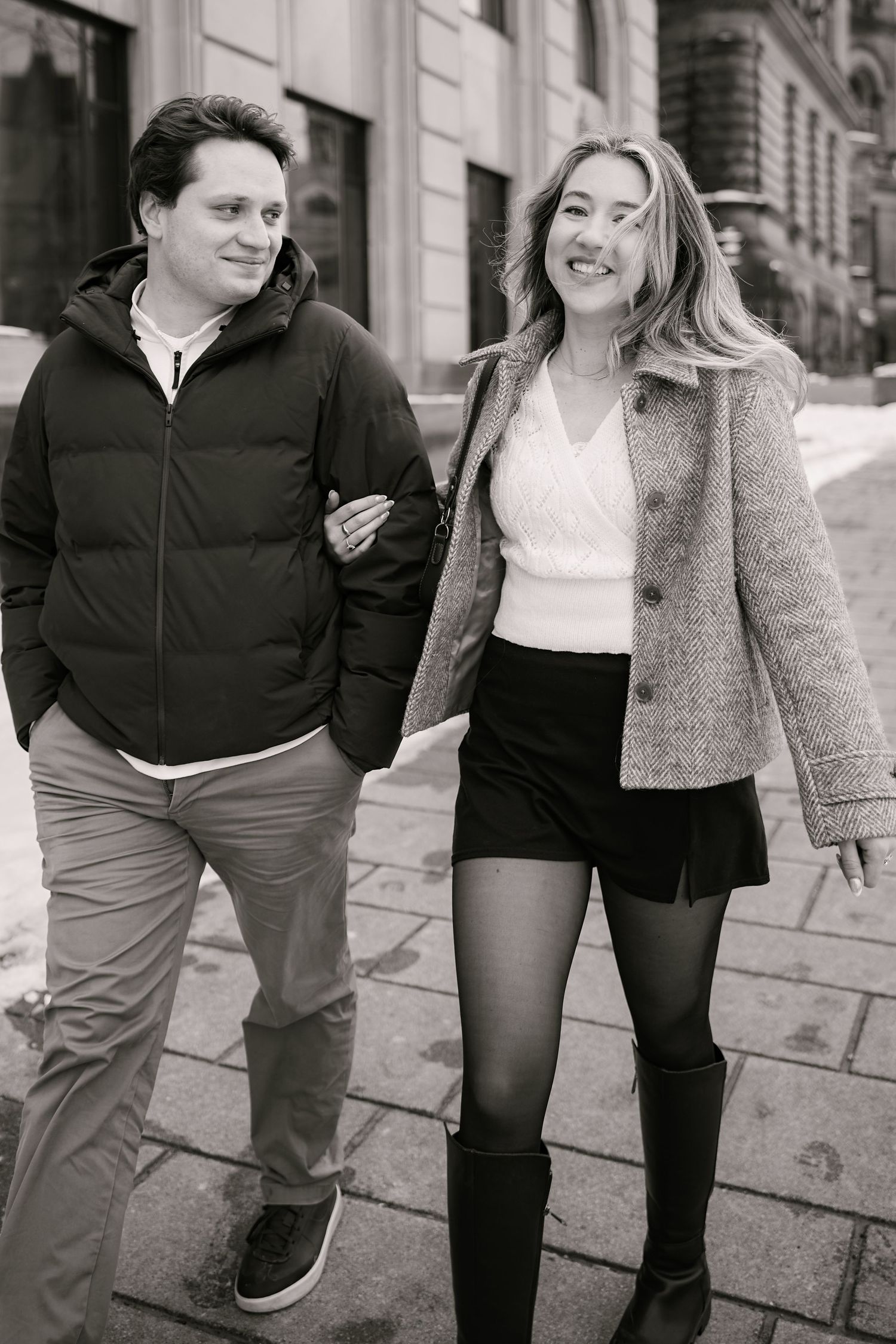 A couple walking arm in arm on a city street, with one in a puffy jacket and the other in a gray coat and shorts.