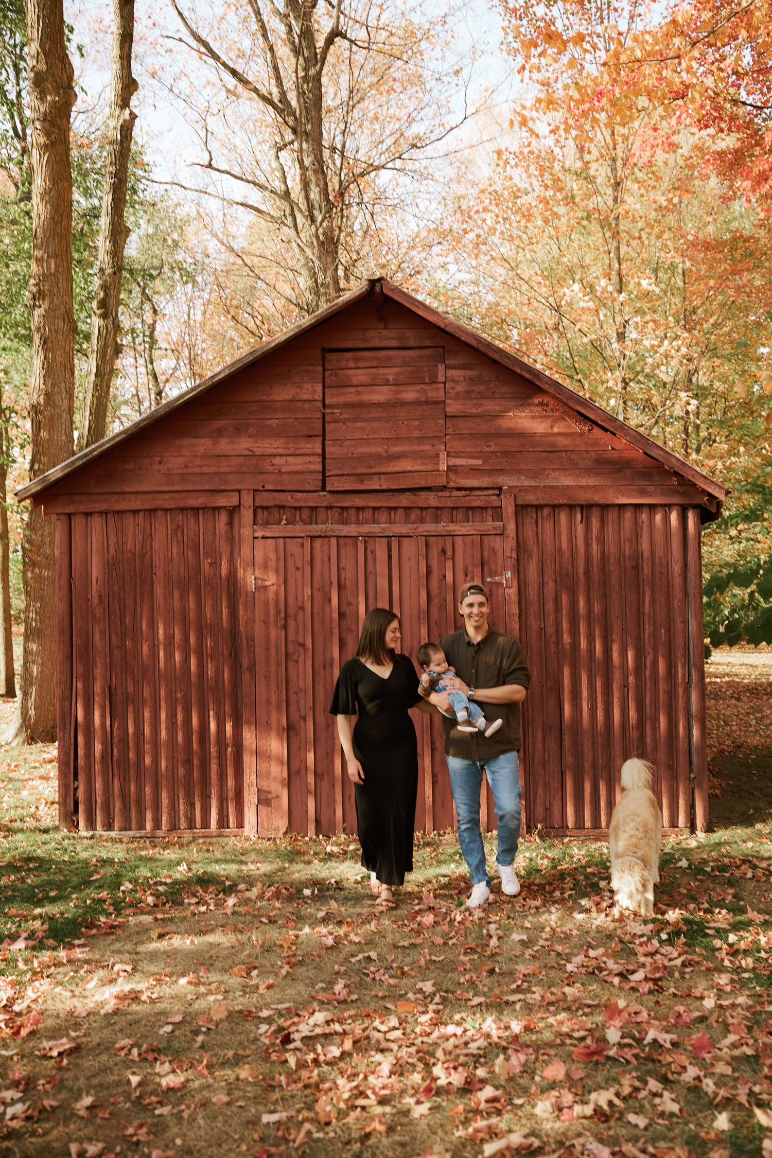 Family photo session by rustic red barn surrounded by autumn trees.