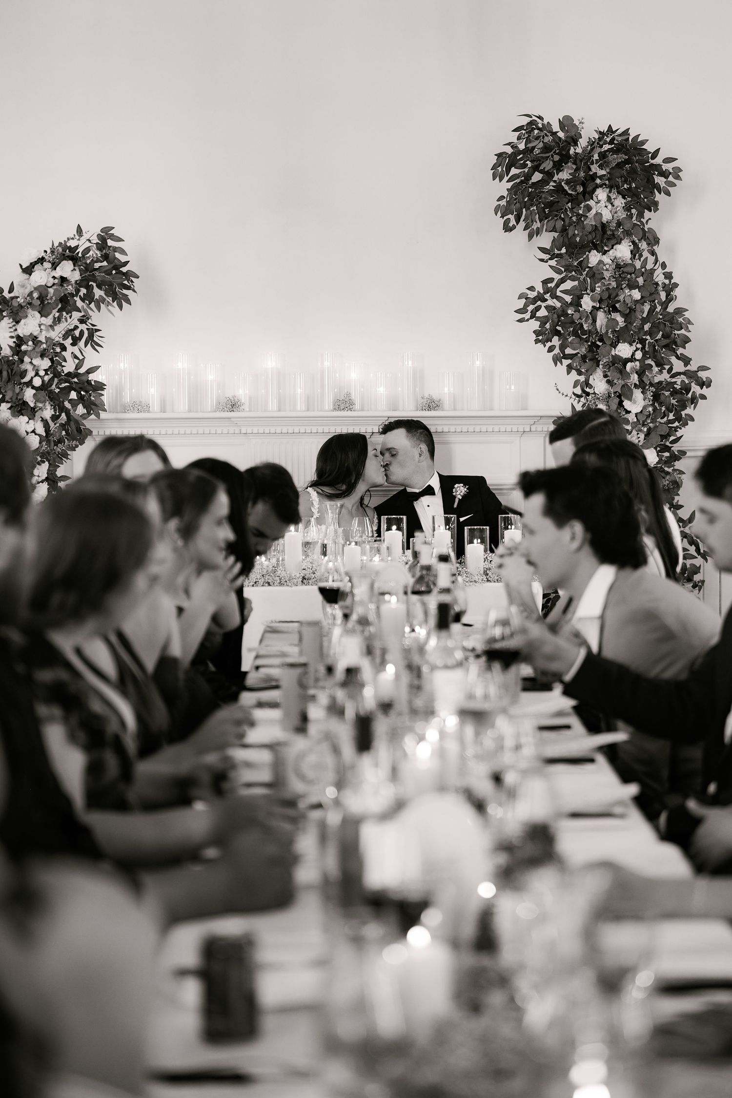 Black and white wedding reception photo of bride and groom kissing at head of long candlelit table surrounded by guests.