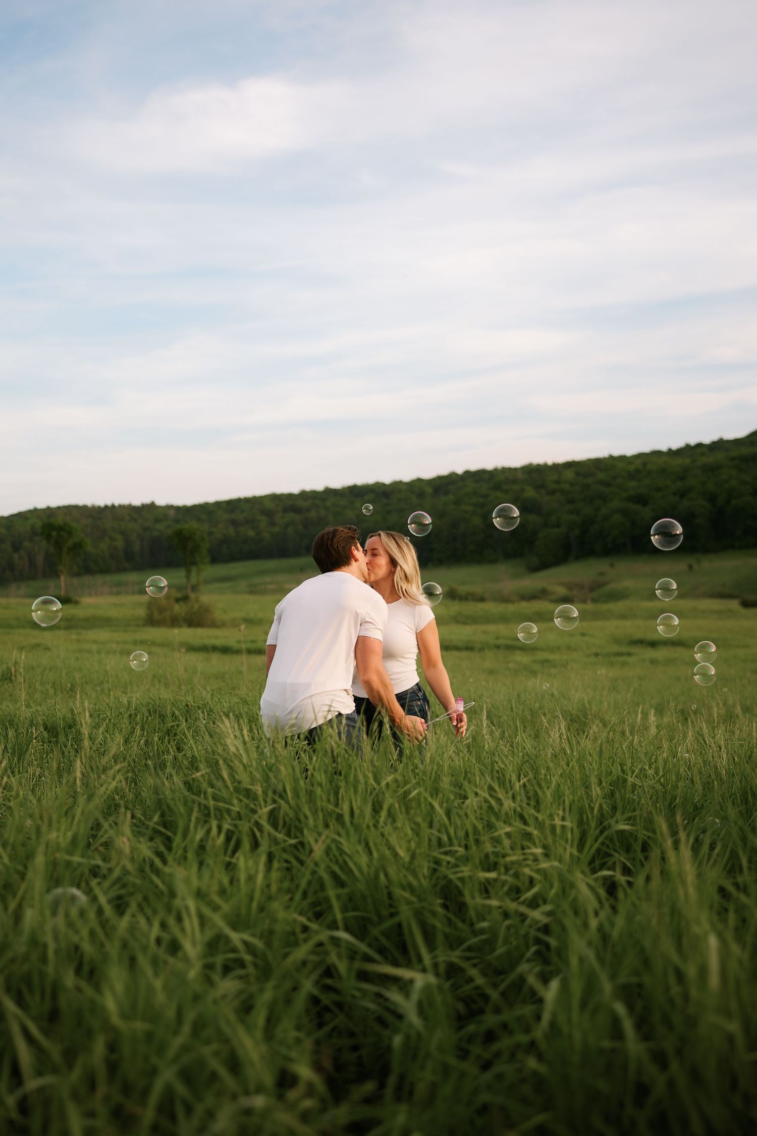 Couple kissing in a lush green field surrounded by floating soap bubbles with rolling hills in the background.