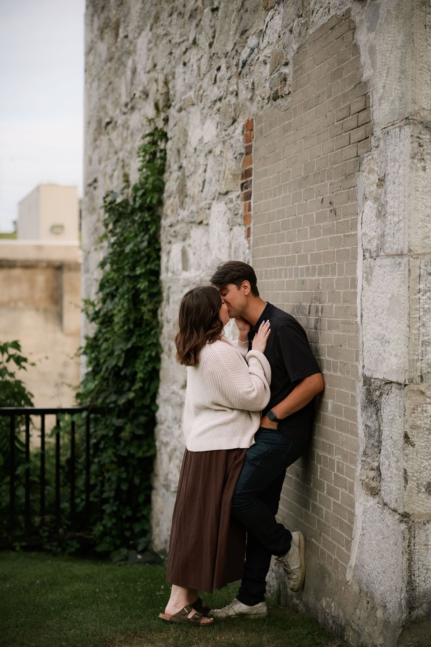 Couple sharing a romantic kiss leaning against a rustic brick wall outdoors, woman in white sweater and brown skirt.
