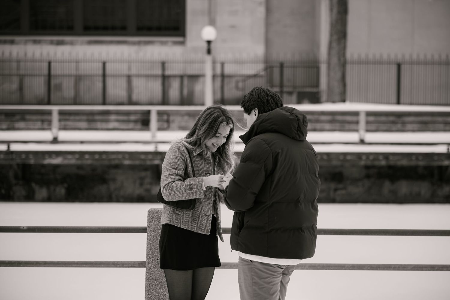 Black and white photo of a couple standing on an ice rink, looking down at something together in winter.
