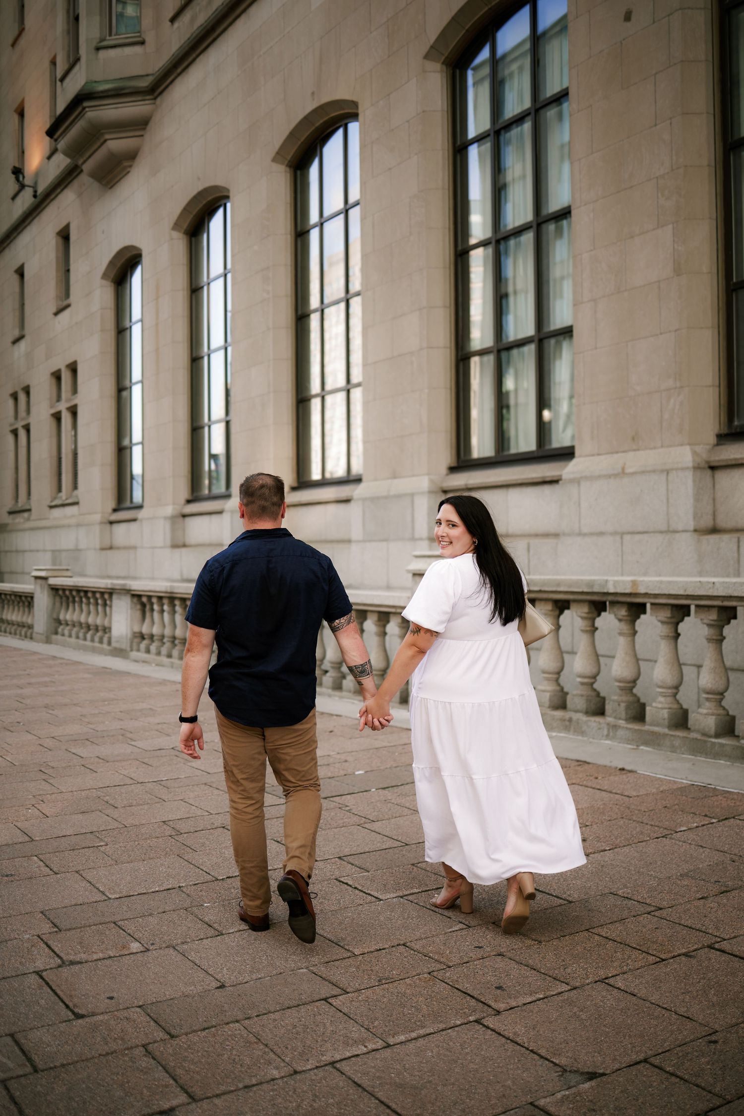 Couple walking hand-in-hand past a grand stone building, woman in white dress glancing back smiling.