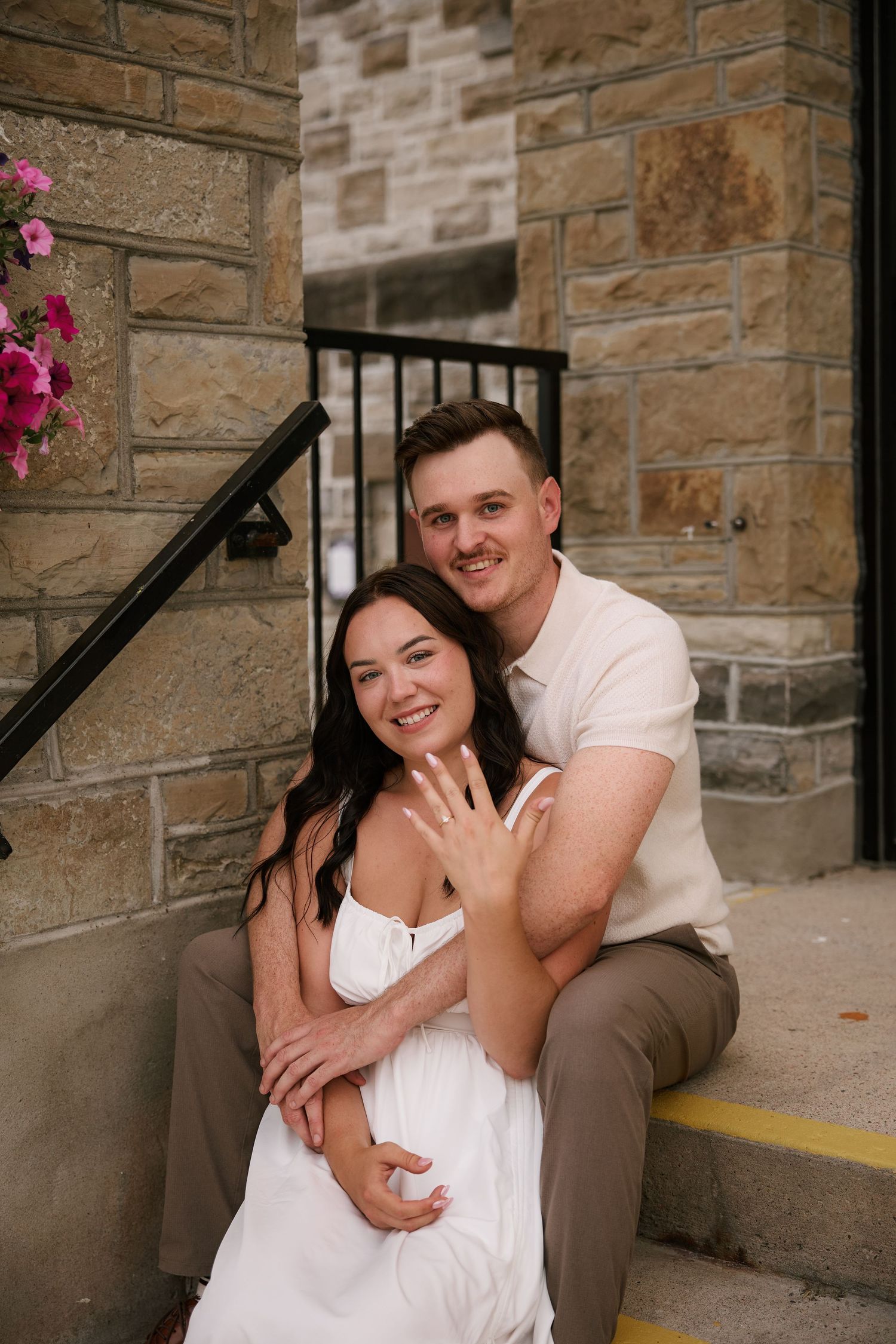 Happy couple sitting on stone steps showing off engagement ring, smiling together in a warm outdoor setting.