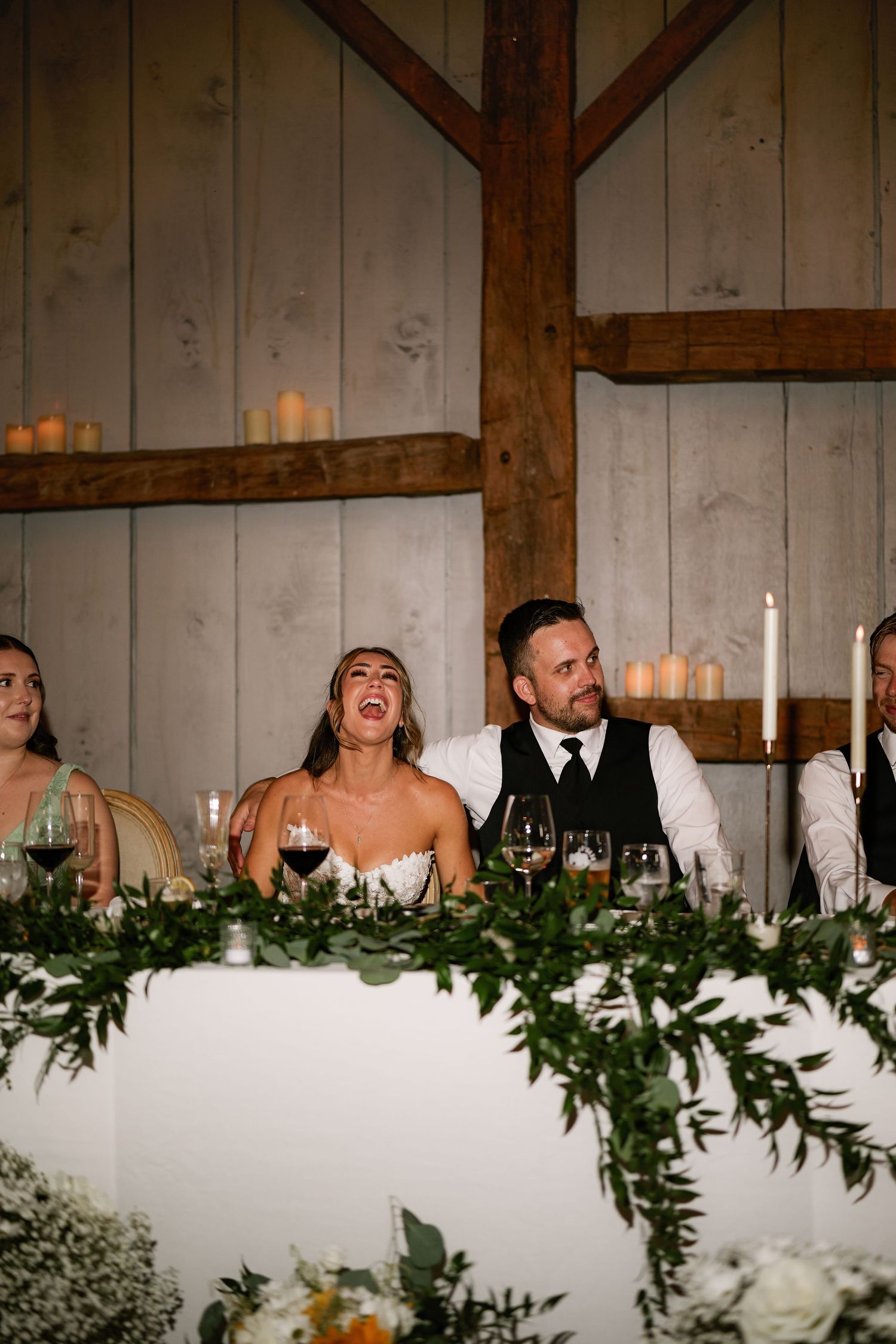 Bride laughing joyfully at wedding reception head table decorated with greenery and candles in rustic barn venue.