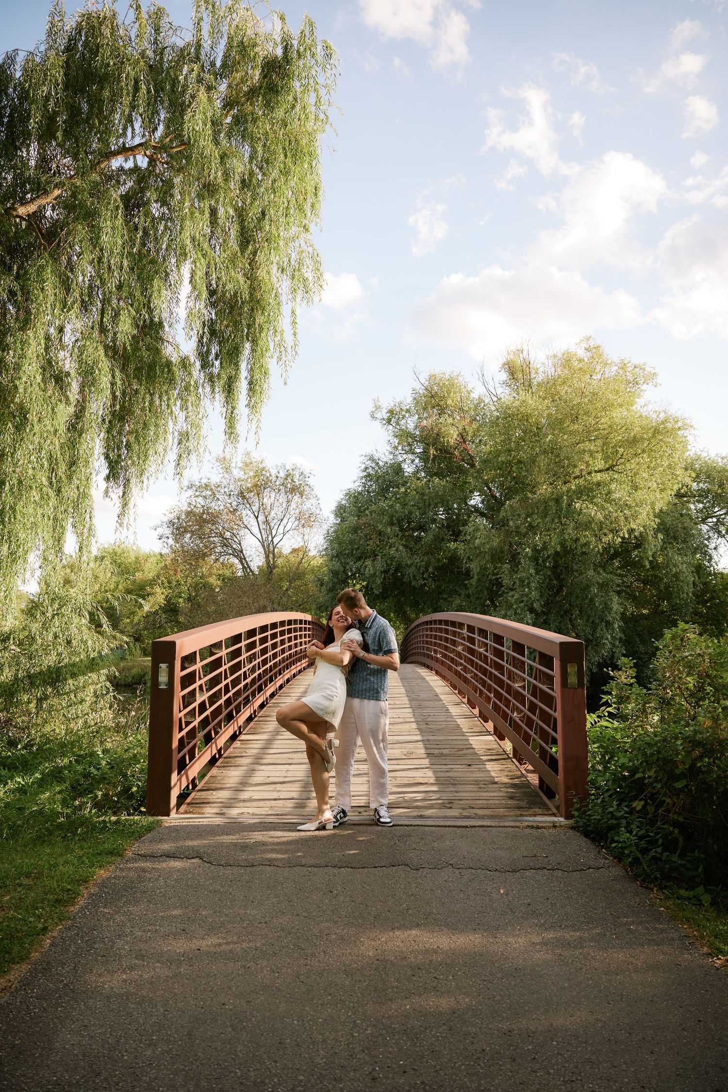 Couple embracing on a wooden bridge surrounded by lush green willow trees in a scenic park setting.