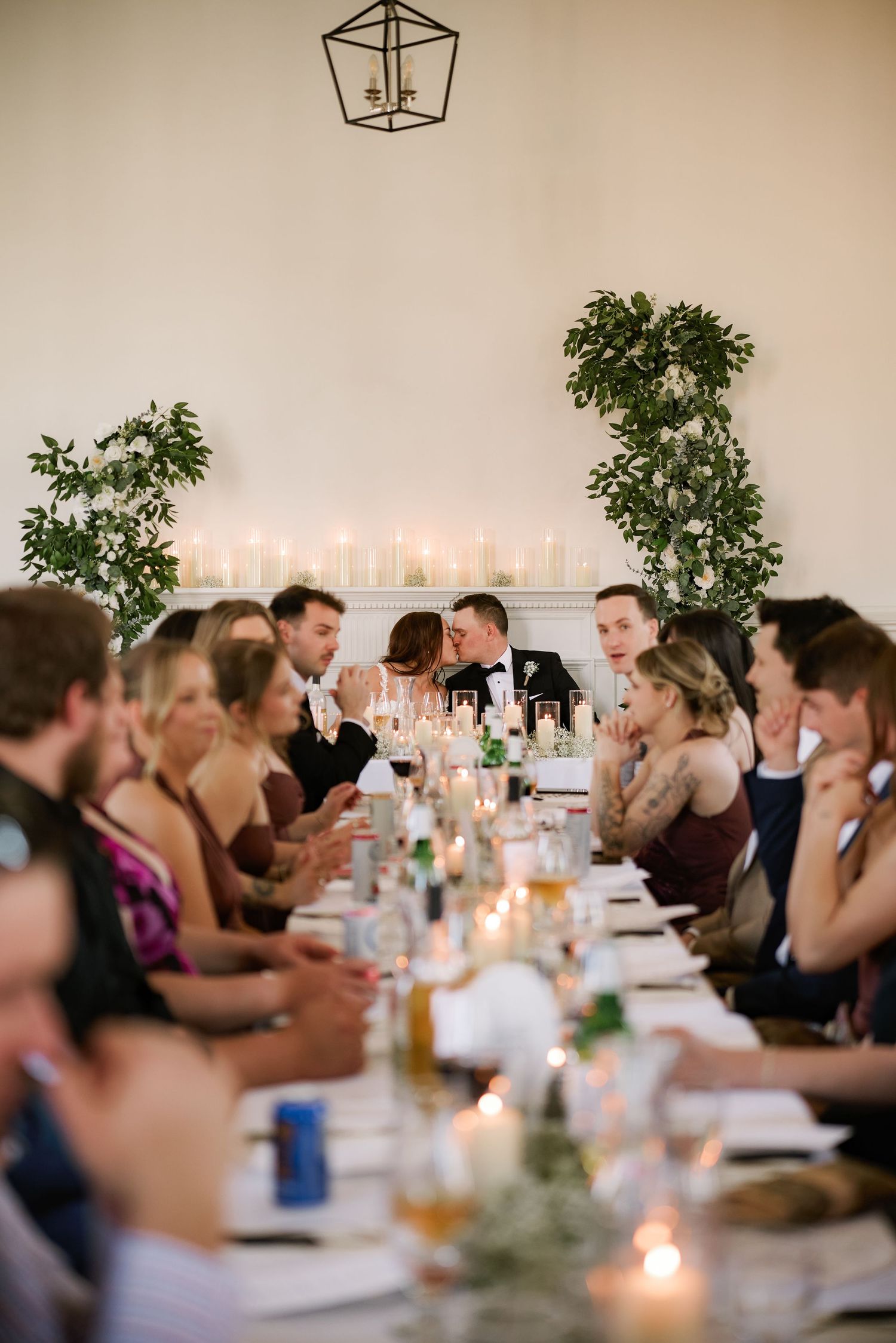 Wedding reception guests seated at long candlelit table while couple shares a kiss at the head table.
