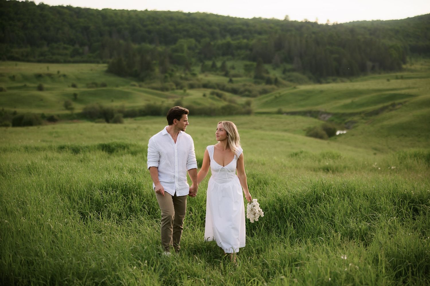 Bride and groom holding hands walking through lush green meadow hills in white wedding attire.