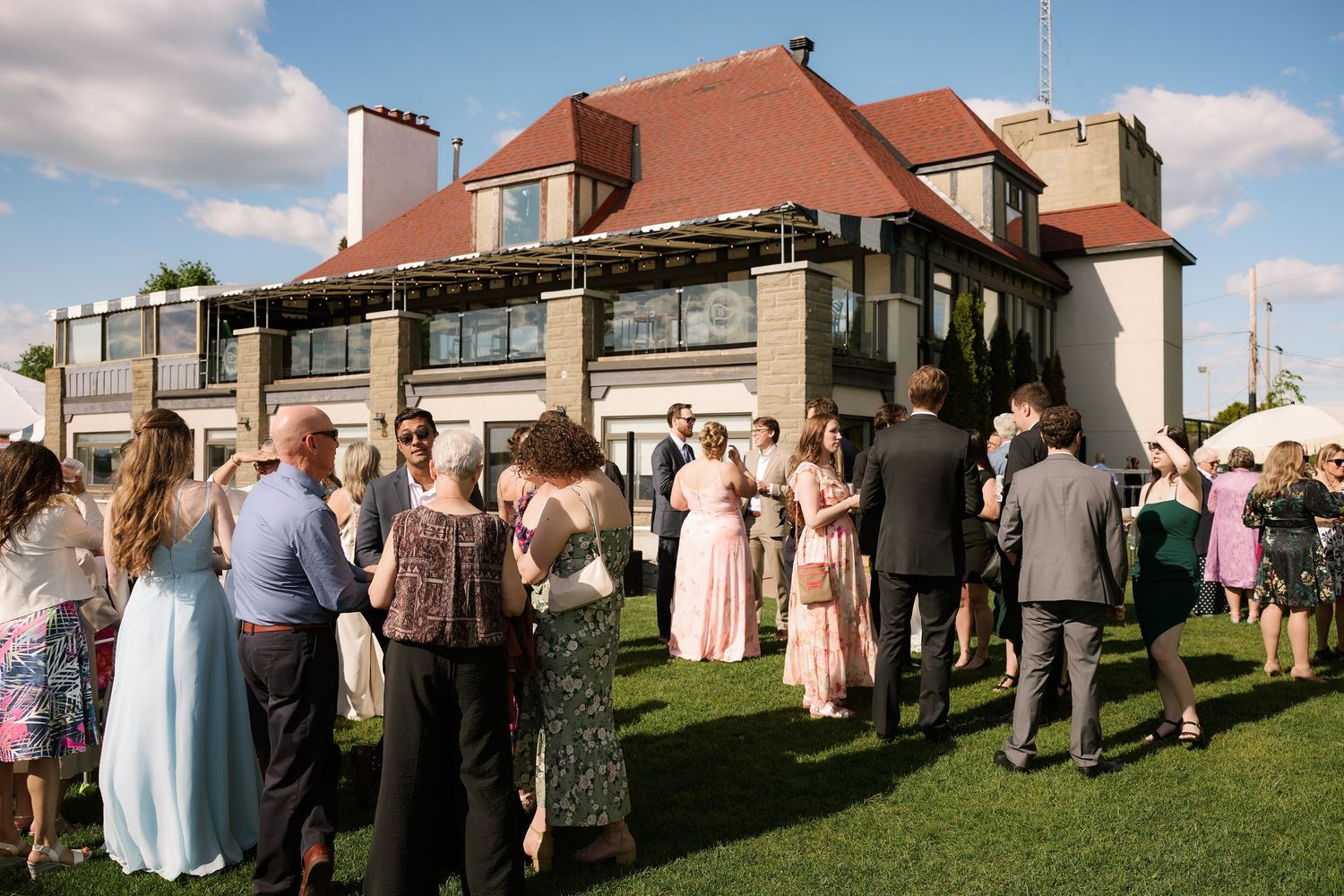 Elegant outdoor wedding reception with guests mingling on green lawn in front of a large brick manor house.