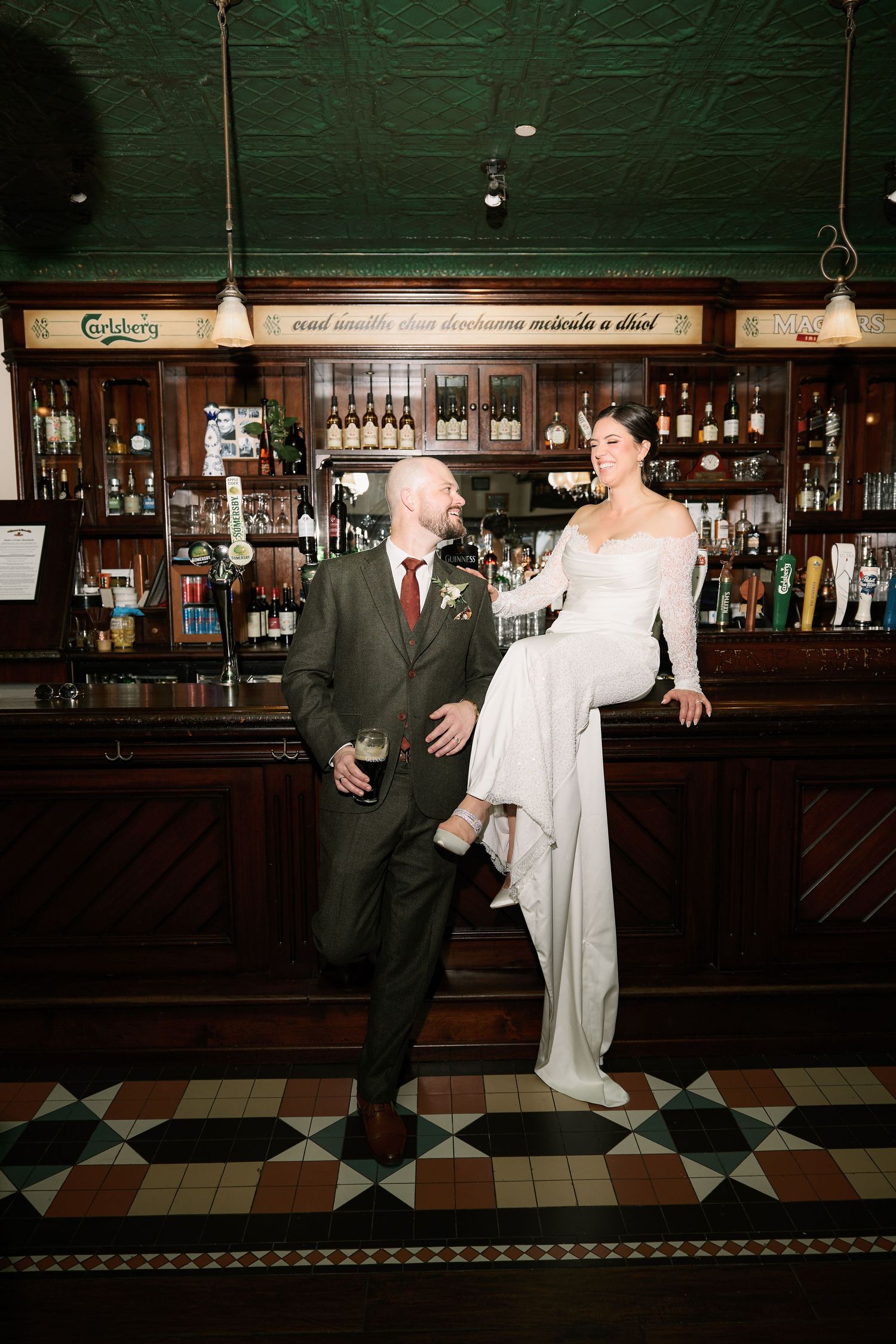 A couple poses playfully at a vintage bar with a classic wooden backdrop and checkerboard floor.