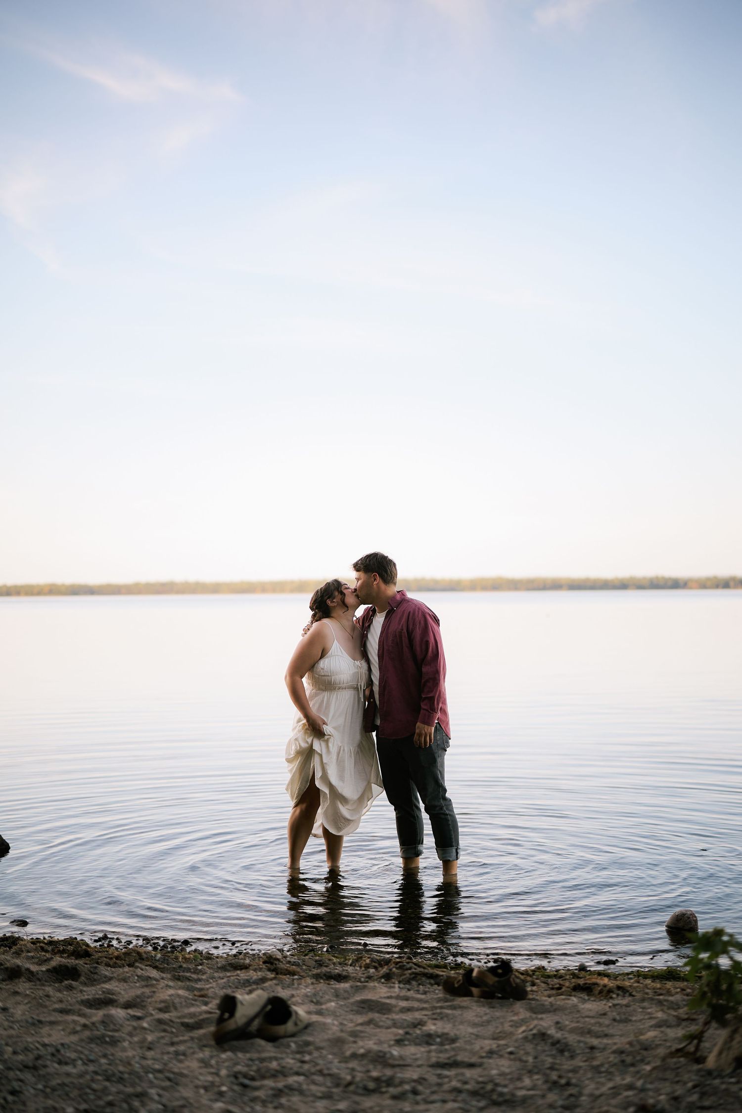 A romantic couple stands together in shallow water at the edge of a serene lake during sunset.