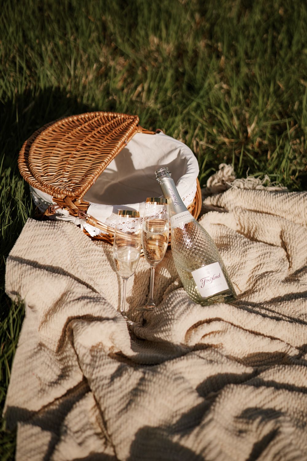 Elegant picnic setup with champagne glasses, bottle, and wicker basket on a linen blanket in green grass.