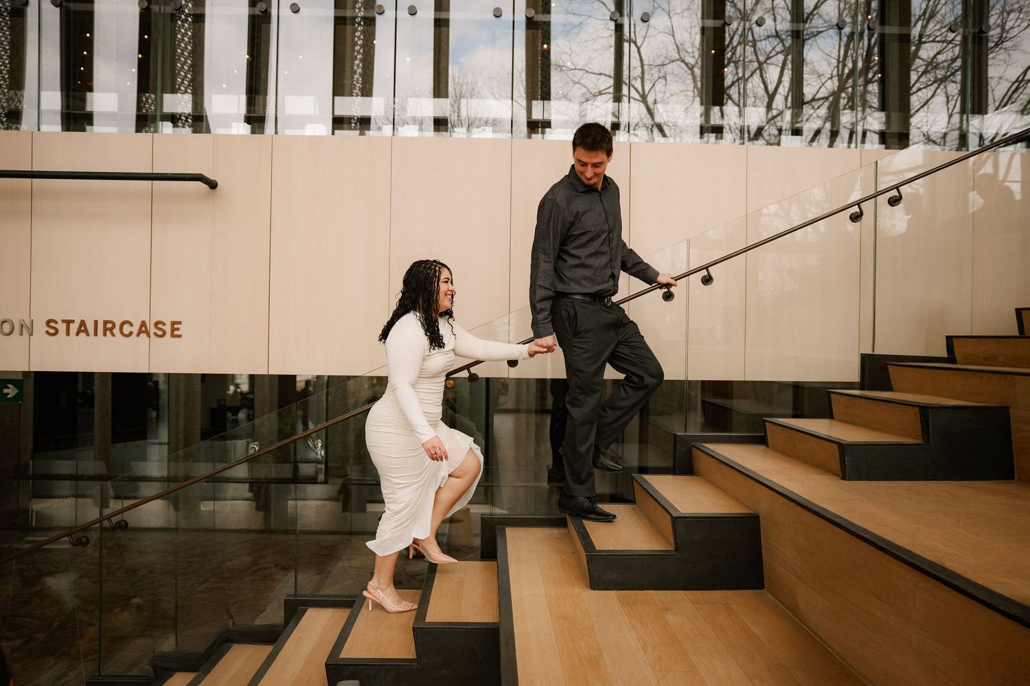 Couple holding hands on modern wooden staircase outside a building, woman in white dress, man in dark outfit.