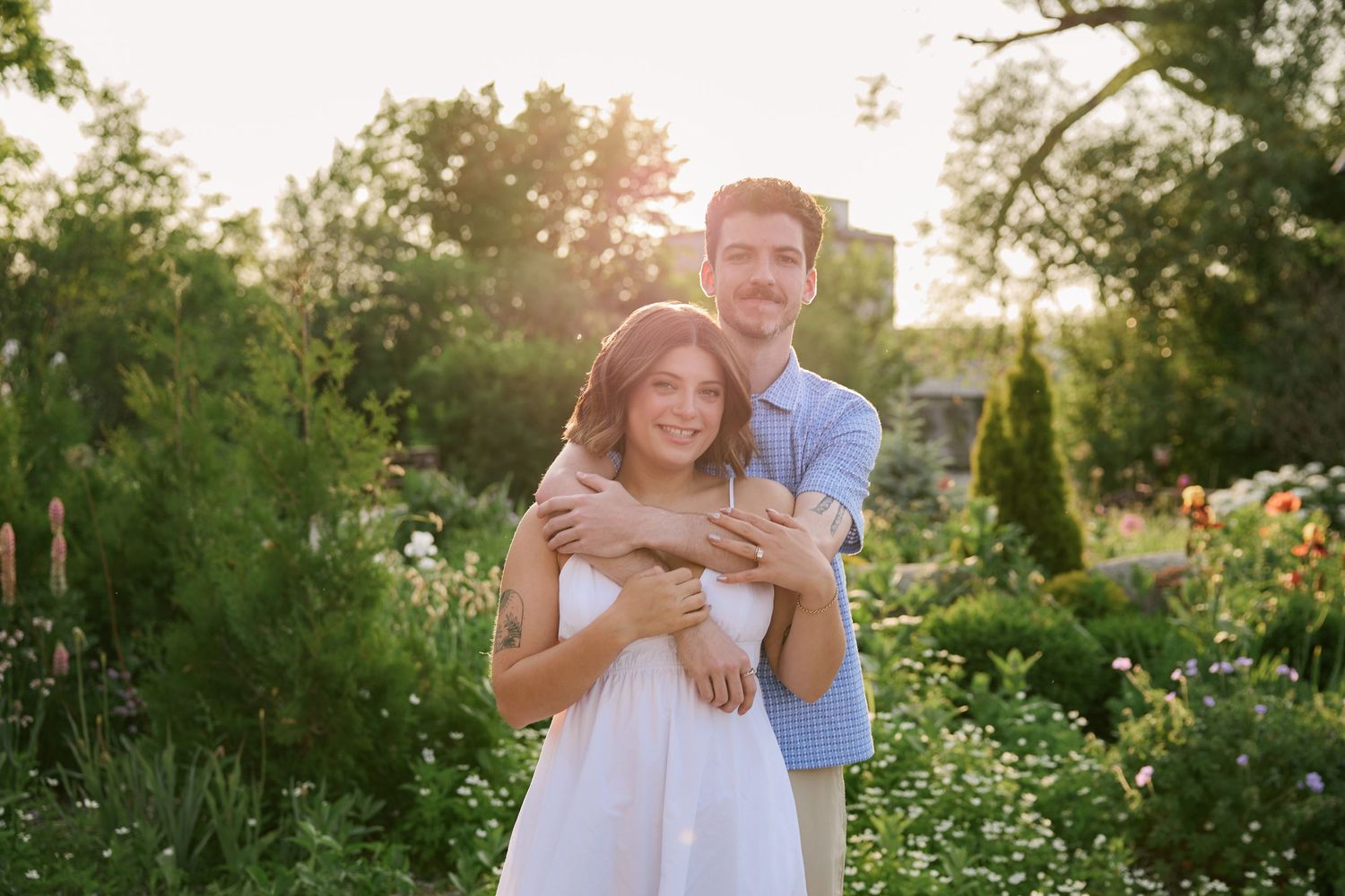 Couple embracing in a lush garden at golden hour, man hugging woman in white dress surrounded by greenery.