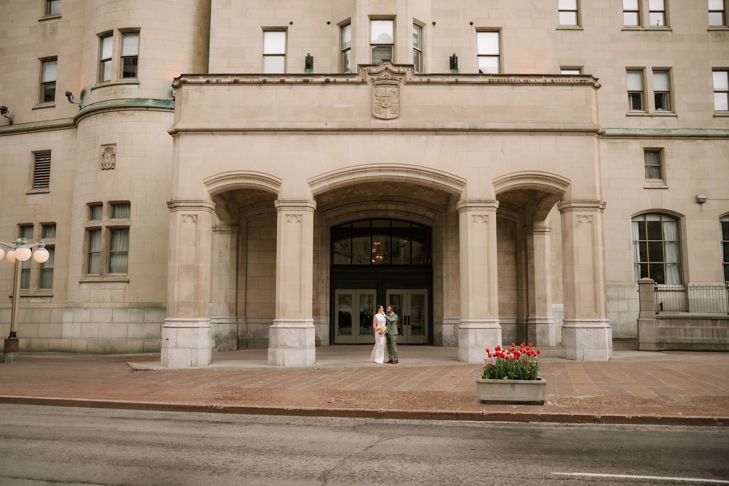 Bride and groom stand beneath grand arched entrance of historic stone building on wedding day.