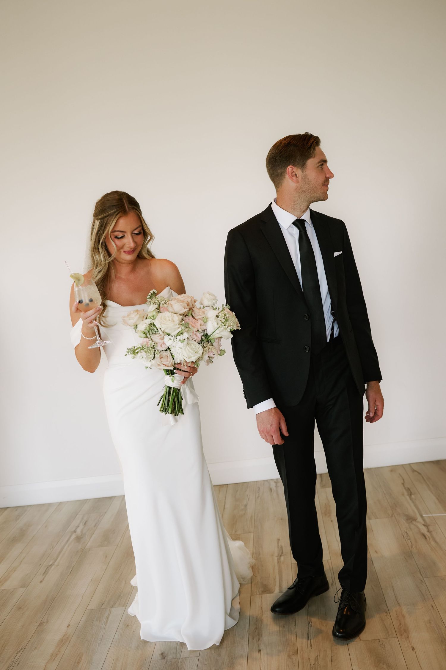 Bride in white gown holding blush bouquet stands beside groom in black tuxedo indoors.