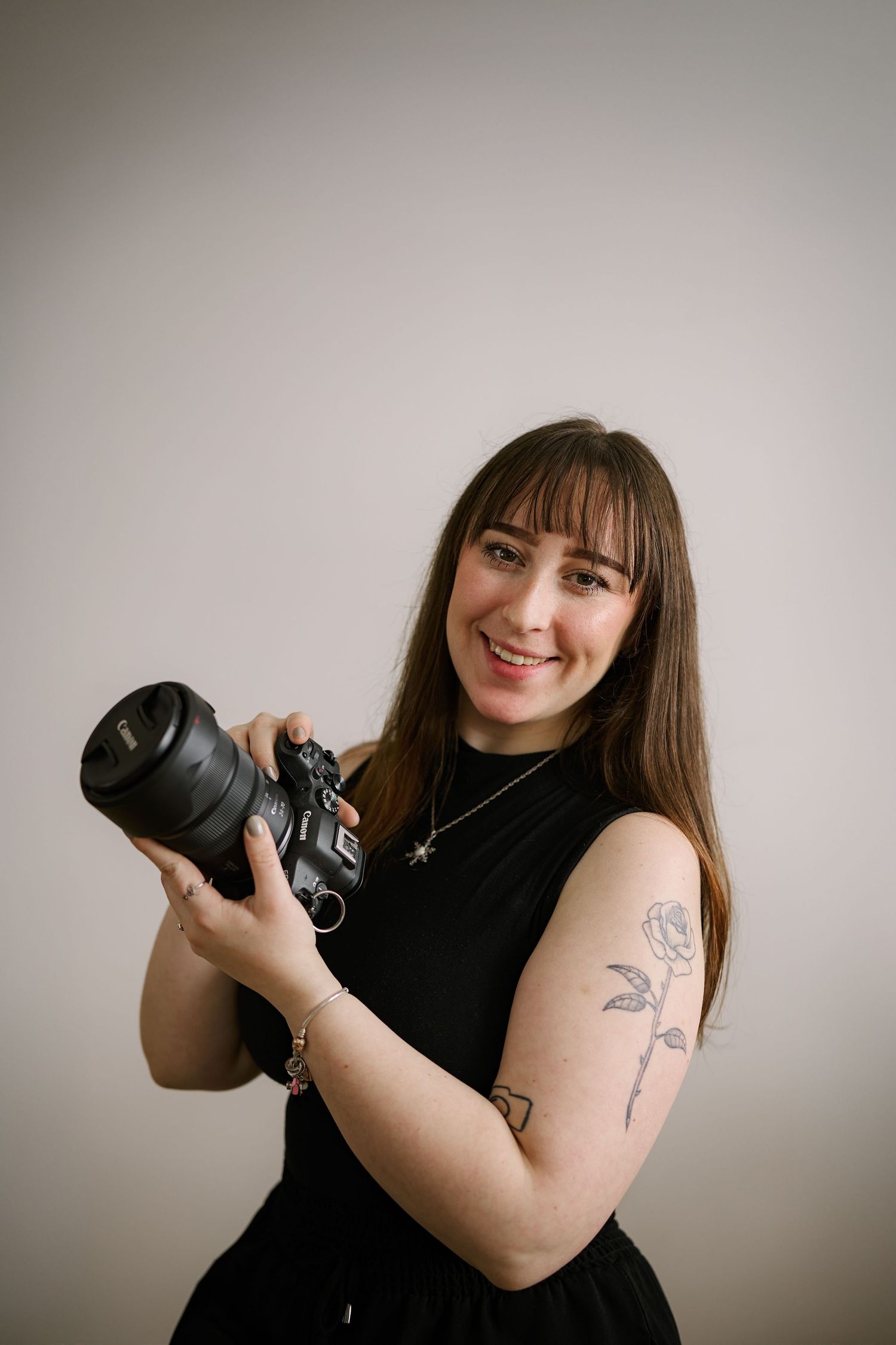 Smiling female photographer holding a DSLR camera, wearing black sleeveless top with rose tattoo on arm.