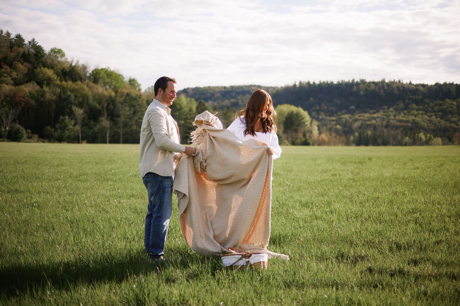 Couple spreading a blanket in a lush green field with rolling hills and trees in the background.