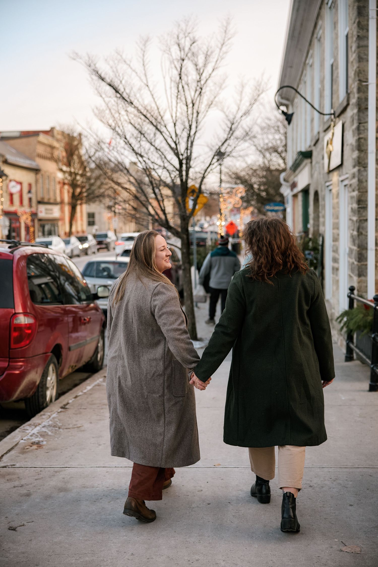 Two women holding hands walk along a charming small-town sidewalk at dusk in matching long coats.