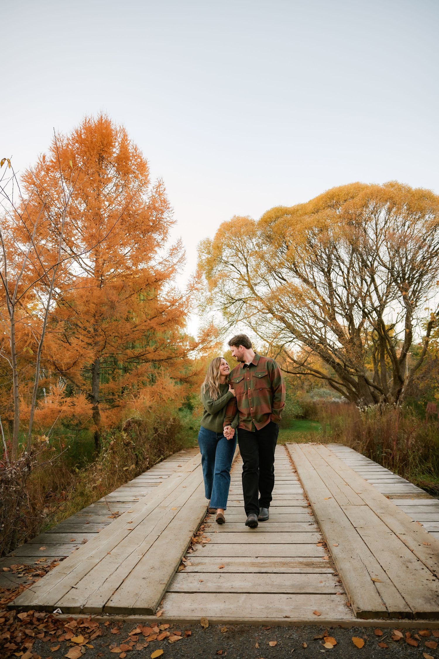 Couple walking hand-in-hand on a wooden bridge surrounded by vibrant autumn foliage in warm orange and gold hues.
