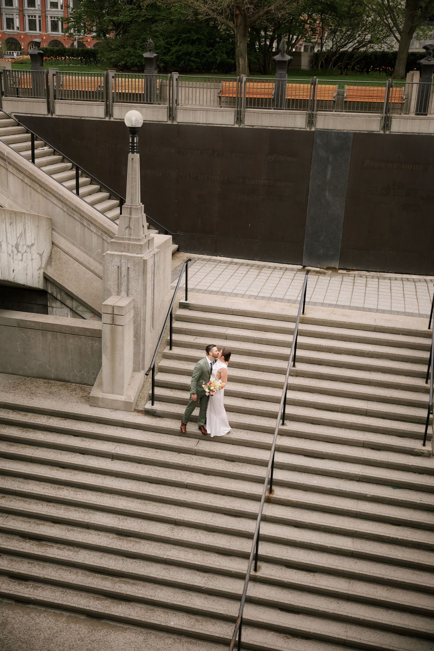 Bride and groom sharing a kiss on wide stone steps at an urban plaza, with elegant staircases and city park in background.