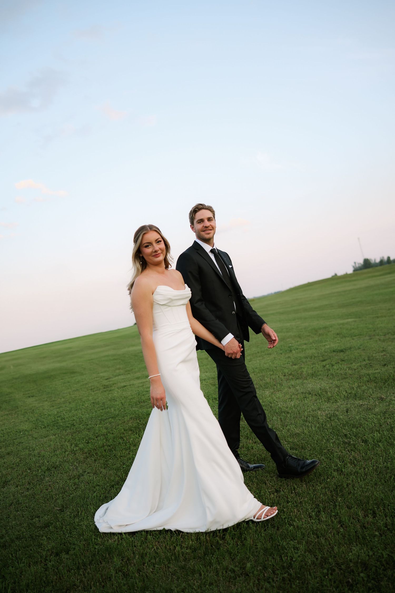 Bride and groom walk hand in hand across green field at dusk in wedding attire.