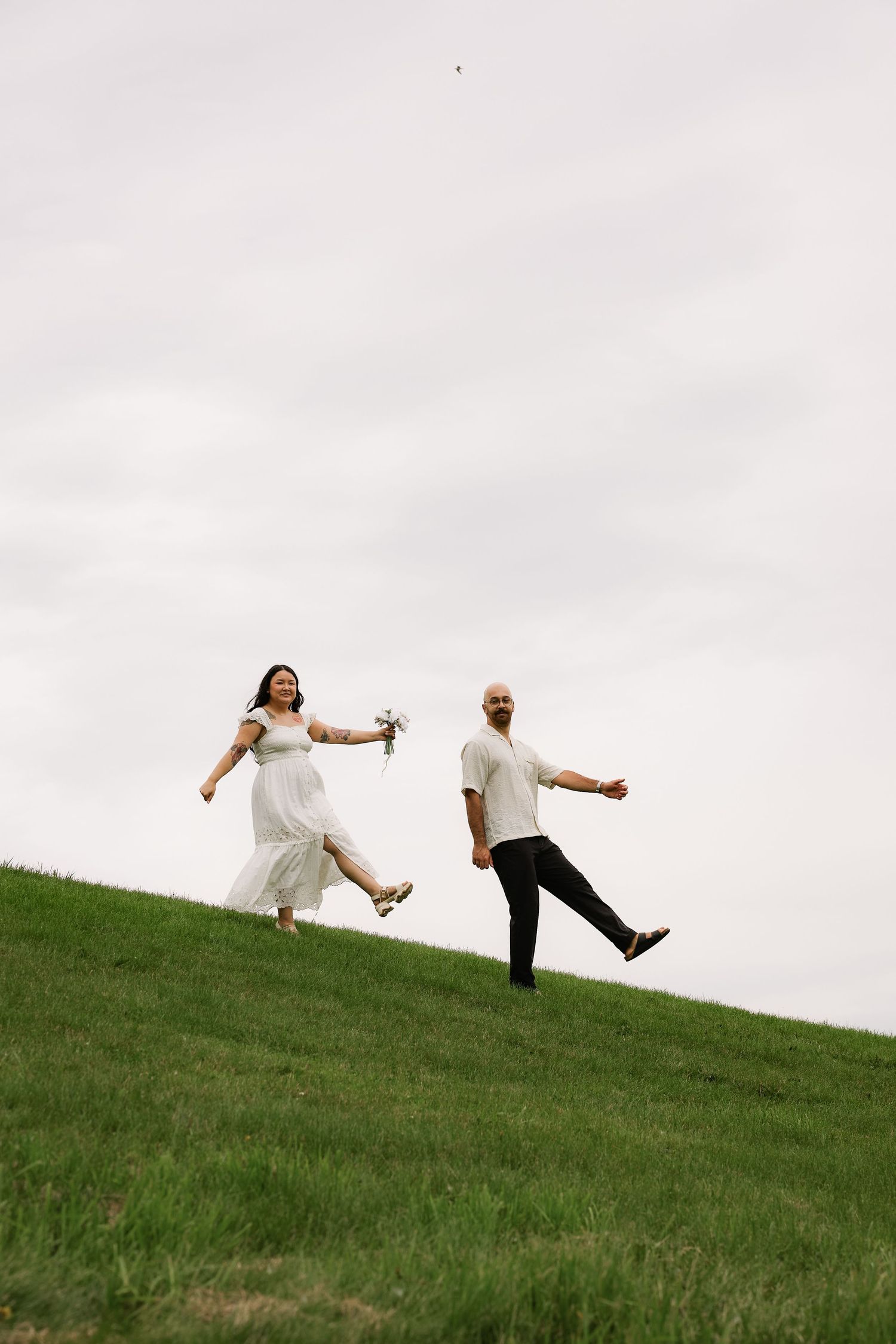 A joyful bride in white dress and groom in black pants run on a green hillside under overcast skies.
