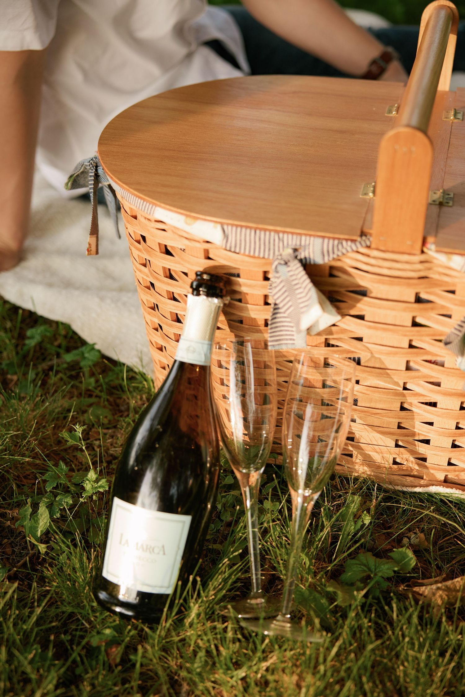 A wicker picnic basket and wine bottle sit on green grass during an outdoor date.