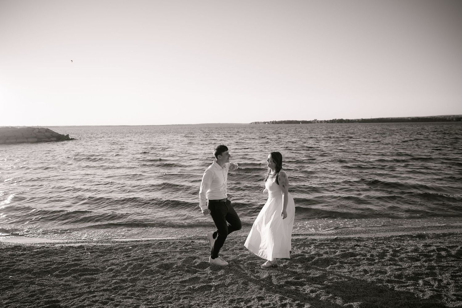 Black and white photo of a bride and groom holding hands walking along a sandy beach shoreline.