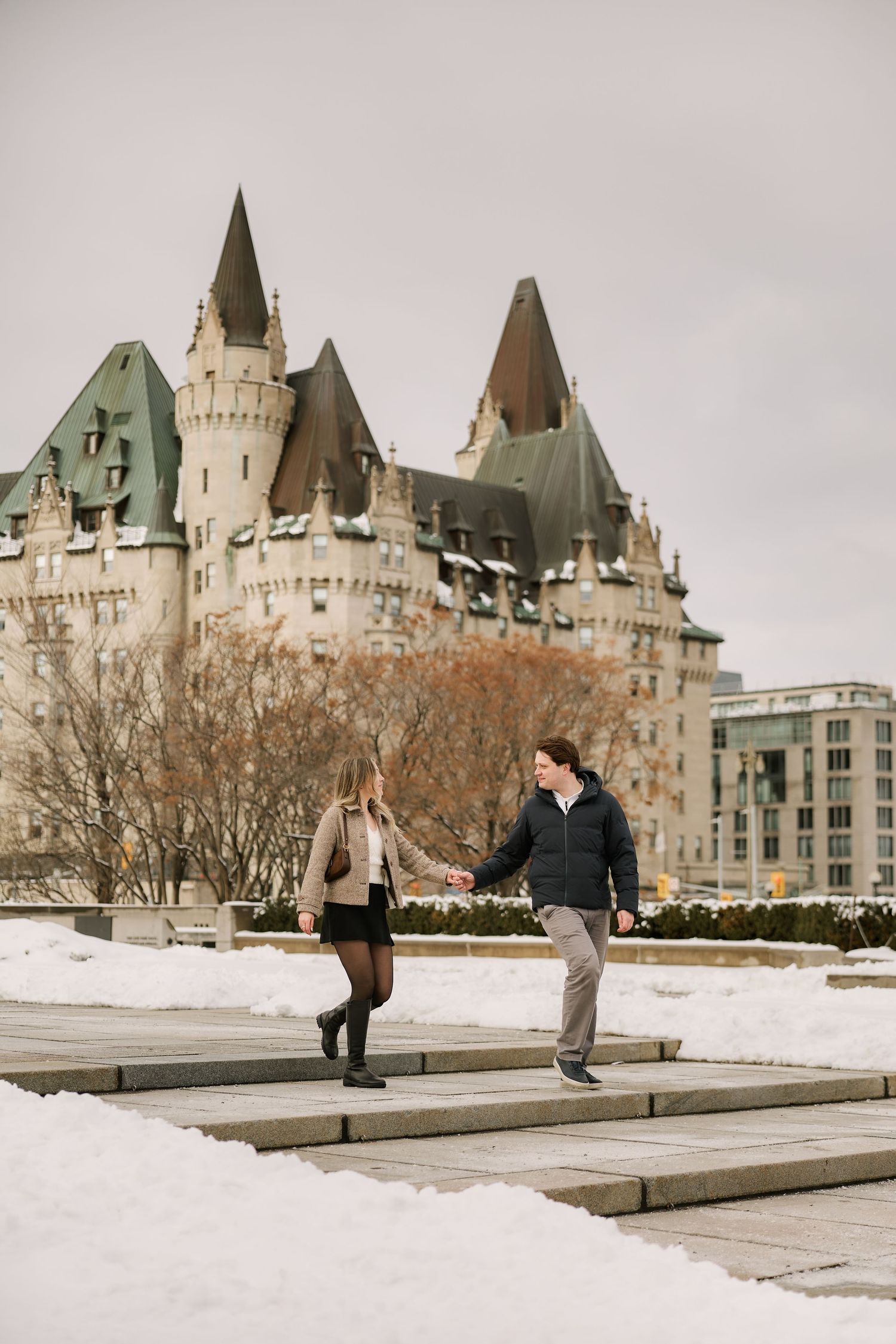 Couple holding hands on snowy steps with Chateau Laurier castle in Ottawa, Canada winter background.